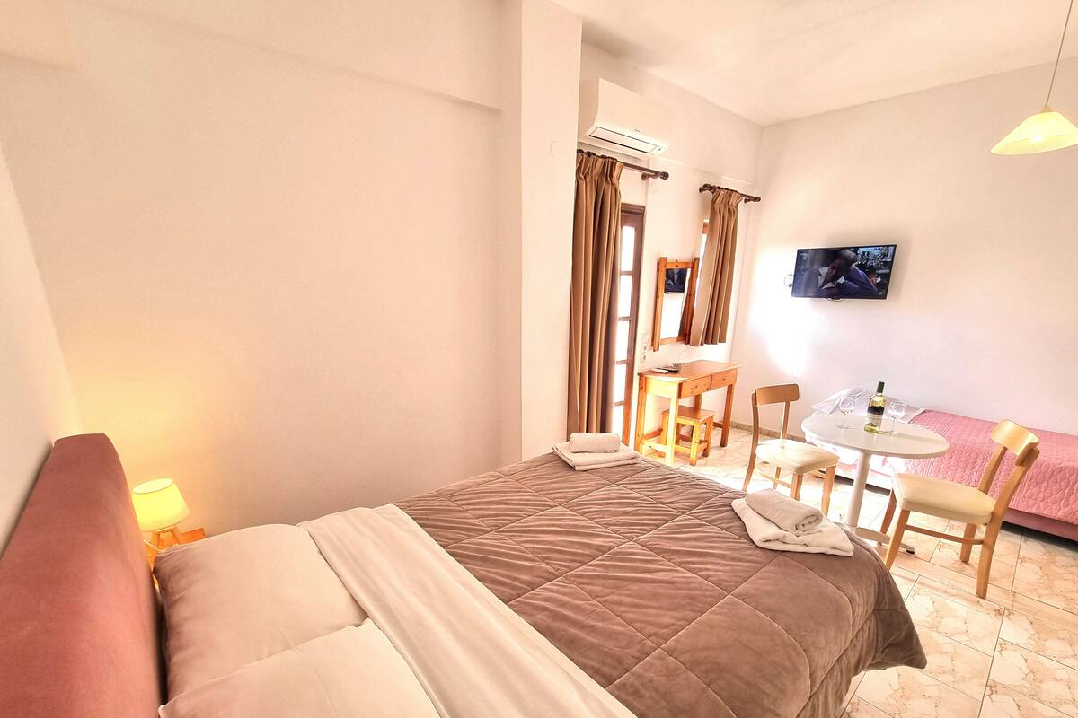 A hotel room with a bed, a desk, a chair, a table with a bottle and glasses, and a TV on the wall.