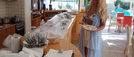 Eine Frau in einem langen Kleid hält einen Teller mit Essen, stehend in einer modernen Küche mit einer Frühstückstheke, Holzschränken und Blick ins Freie.