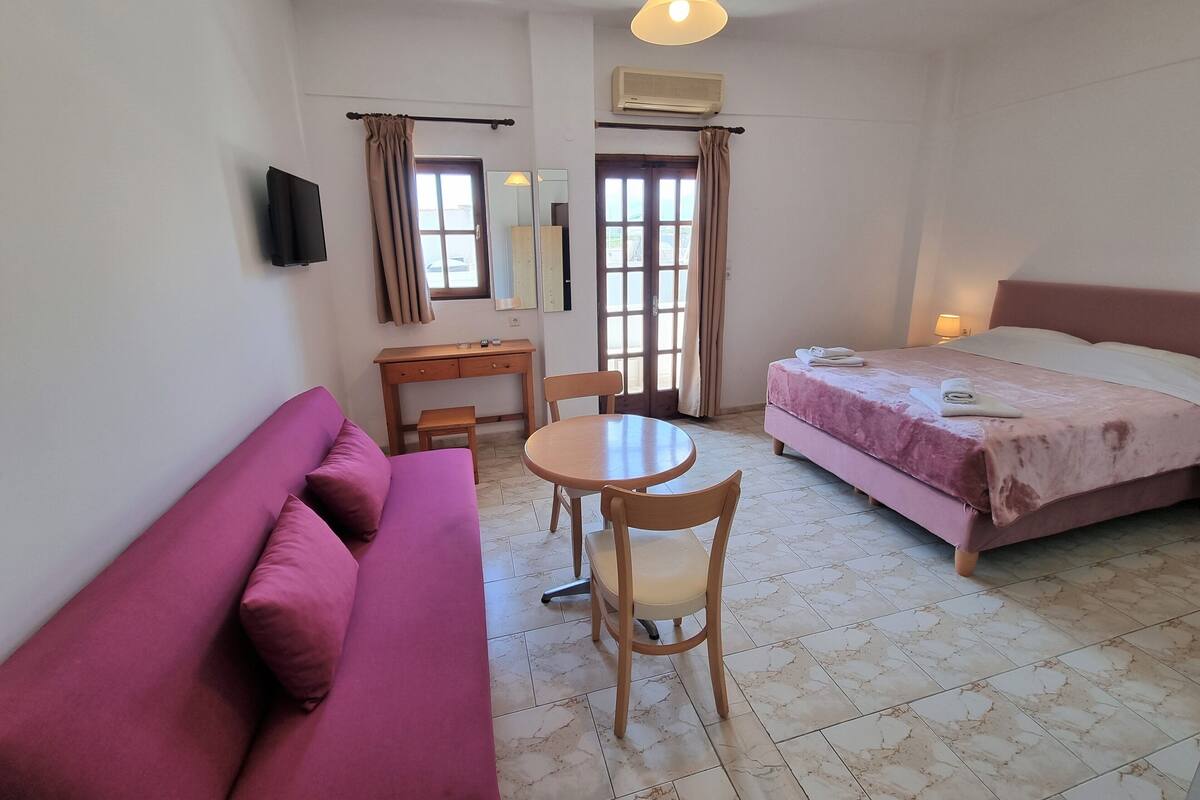 A hotel room with a bed, a sofa, a desk, a chair, a table, a lamp, and an air conditioning unit.