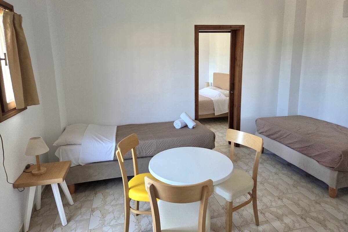A small, clean room with two single beds, a round table with chairs, a small table with a lamp, and a door leading to another room.