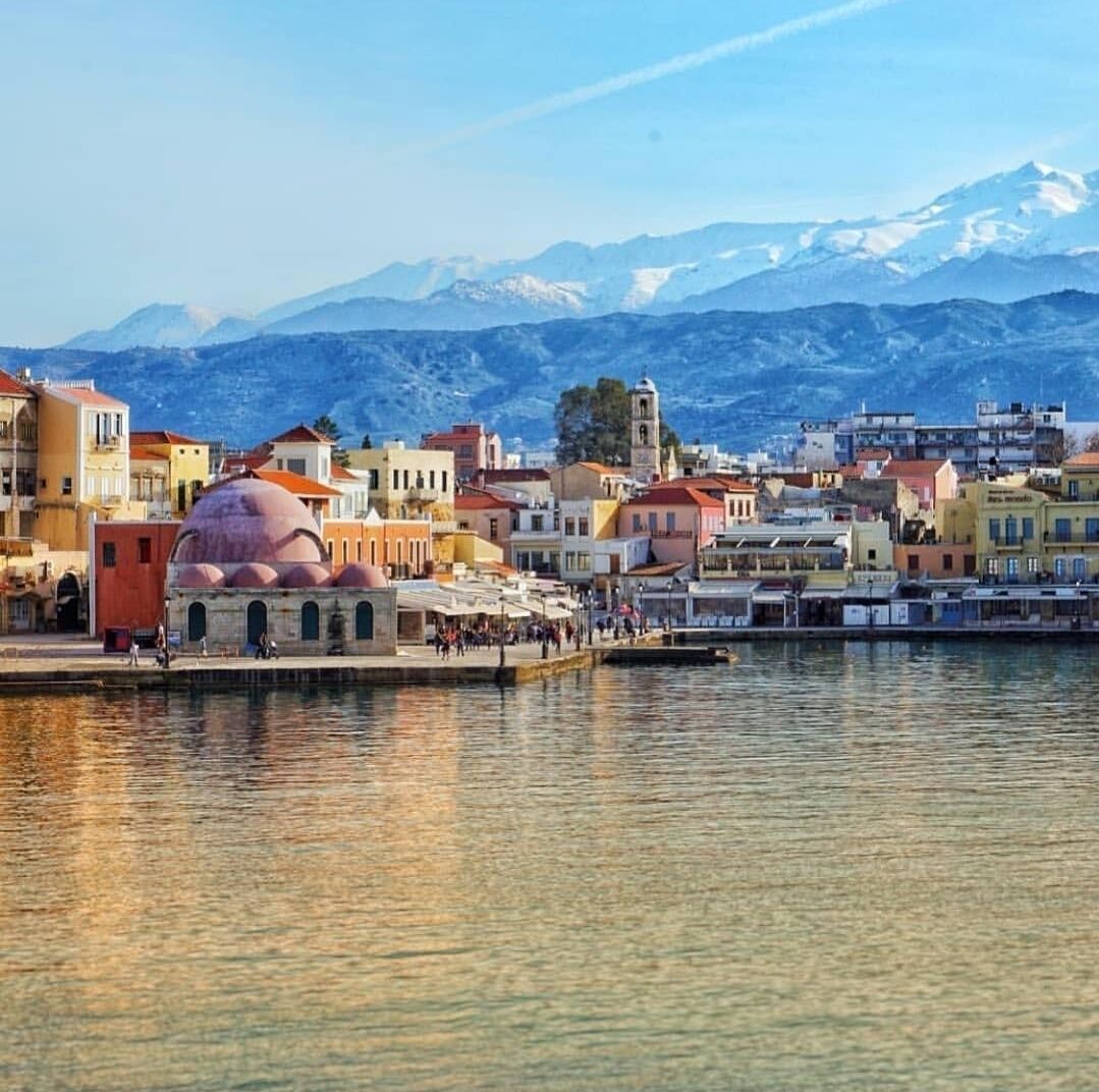 A coastal town with colorful buildings, a prominent dome structure, and snow-capped mountains in the background.