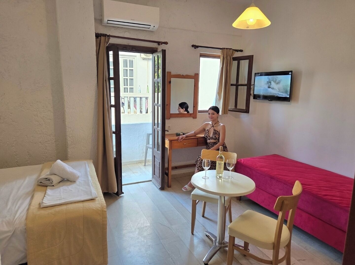 Ett hotellrum med en säng, ett skrivbord, en stol, ett bord med glas, en tv och en balkong.