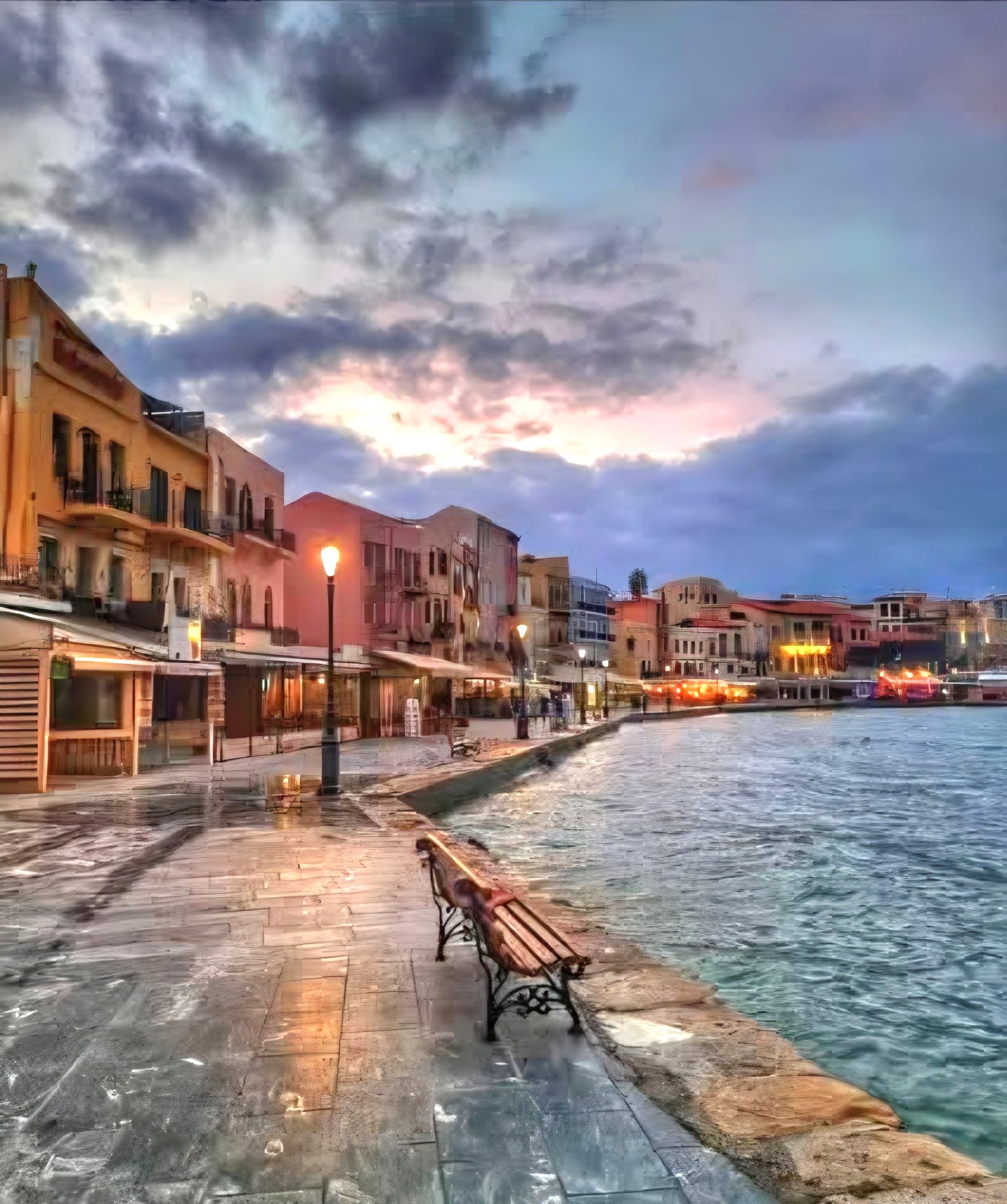 A waterfront promenade with benches and street lamps, buildings lining the water, and a sunset in the background.
