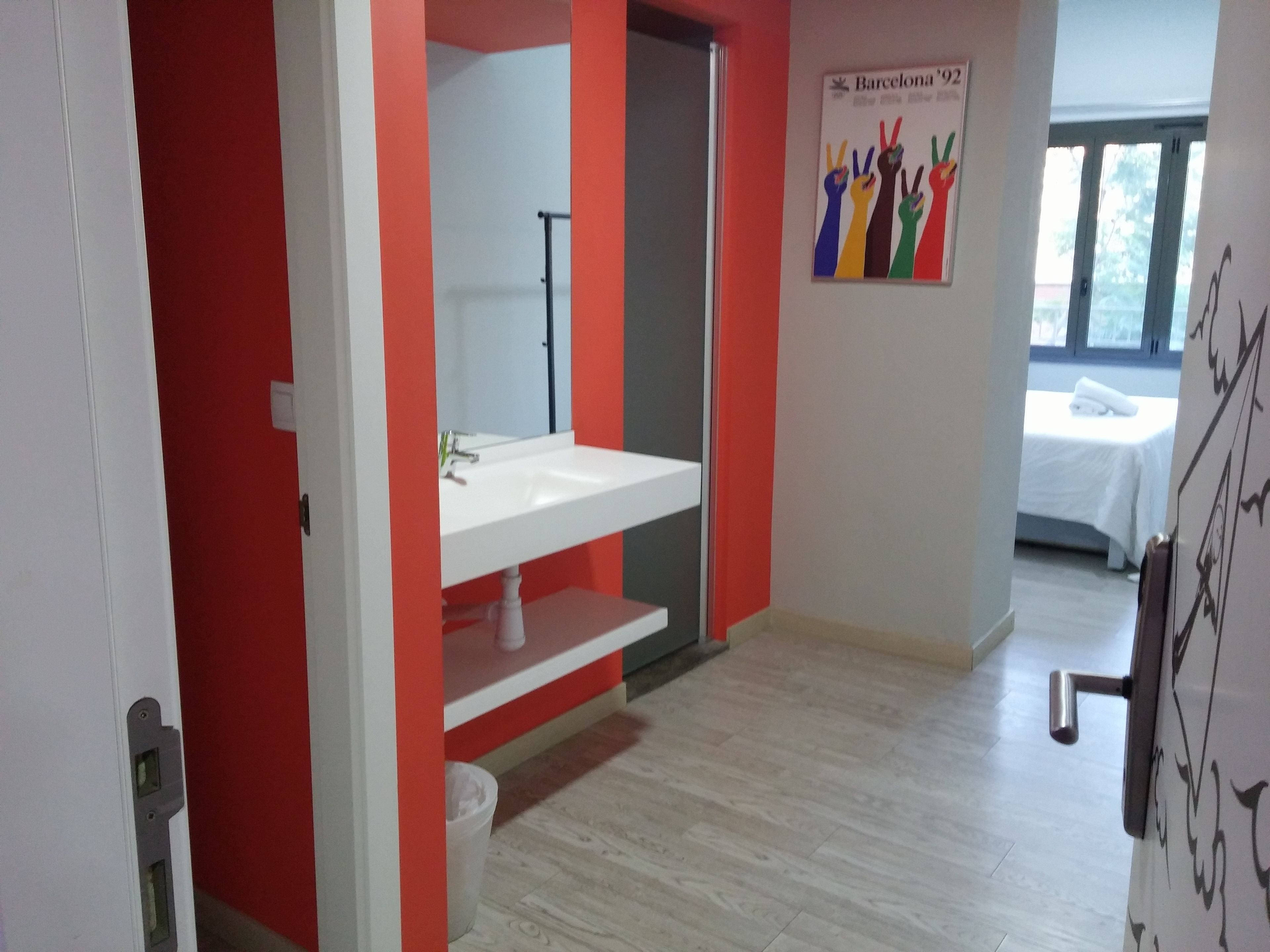 A hotel bathroom with a white sink, red walls, and a poster of the Barcelona '92 Olympics.