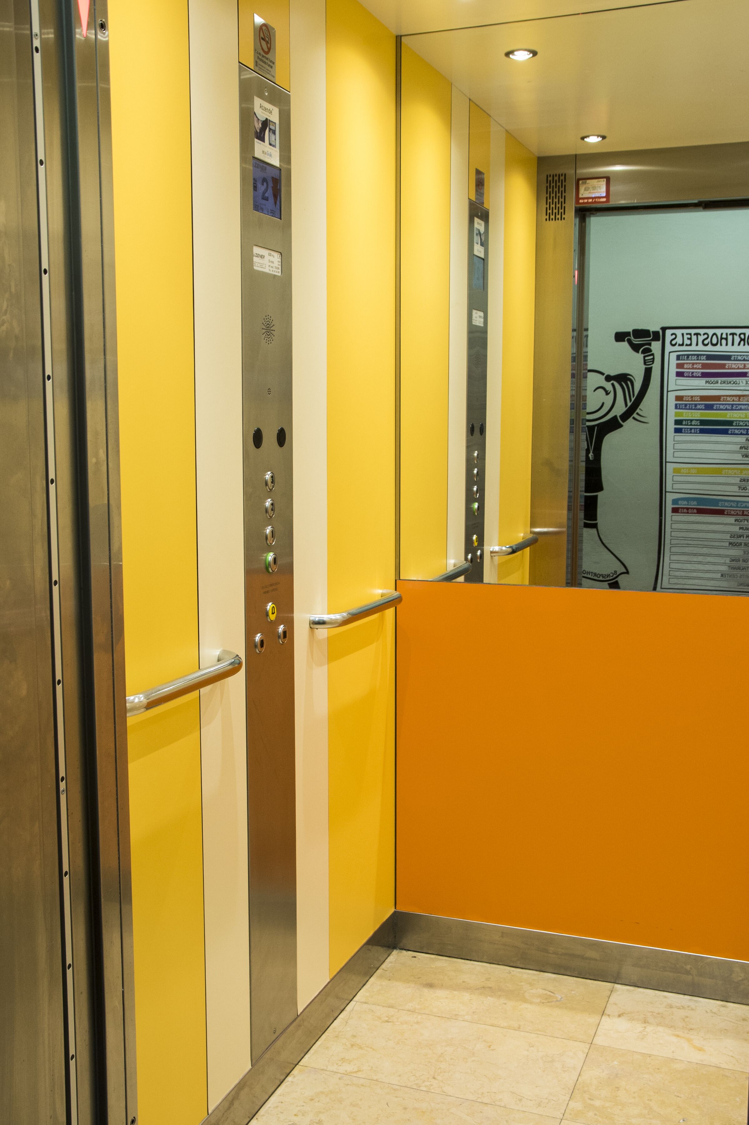 Un ascensor con paredes amarillo blanco, un pasamanos de metal y un piso con baldosas color crema.