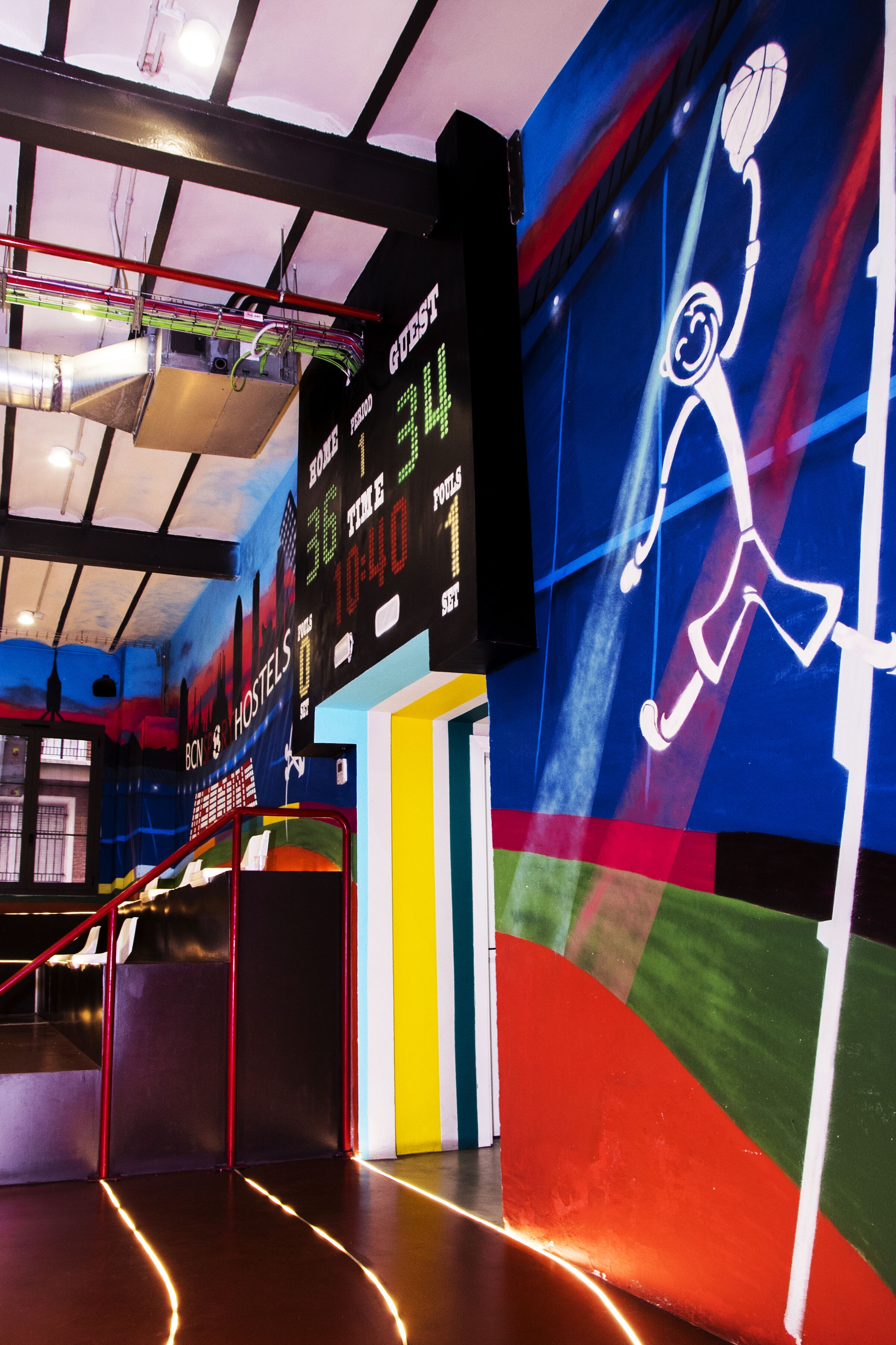 A brightly lit indoor space with a basketball court, a digital scoreboard, and a colorful mural.