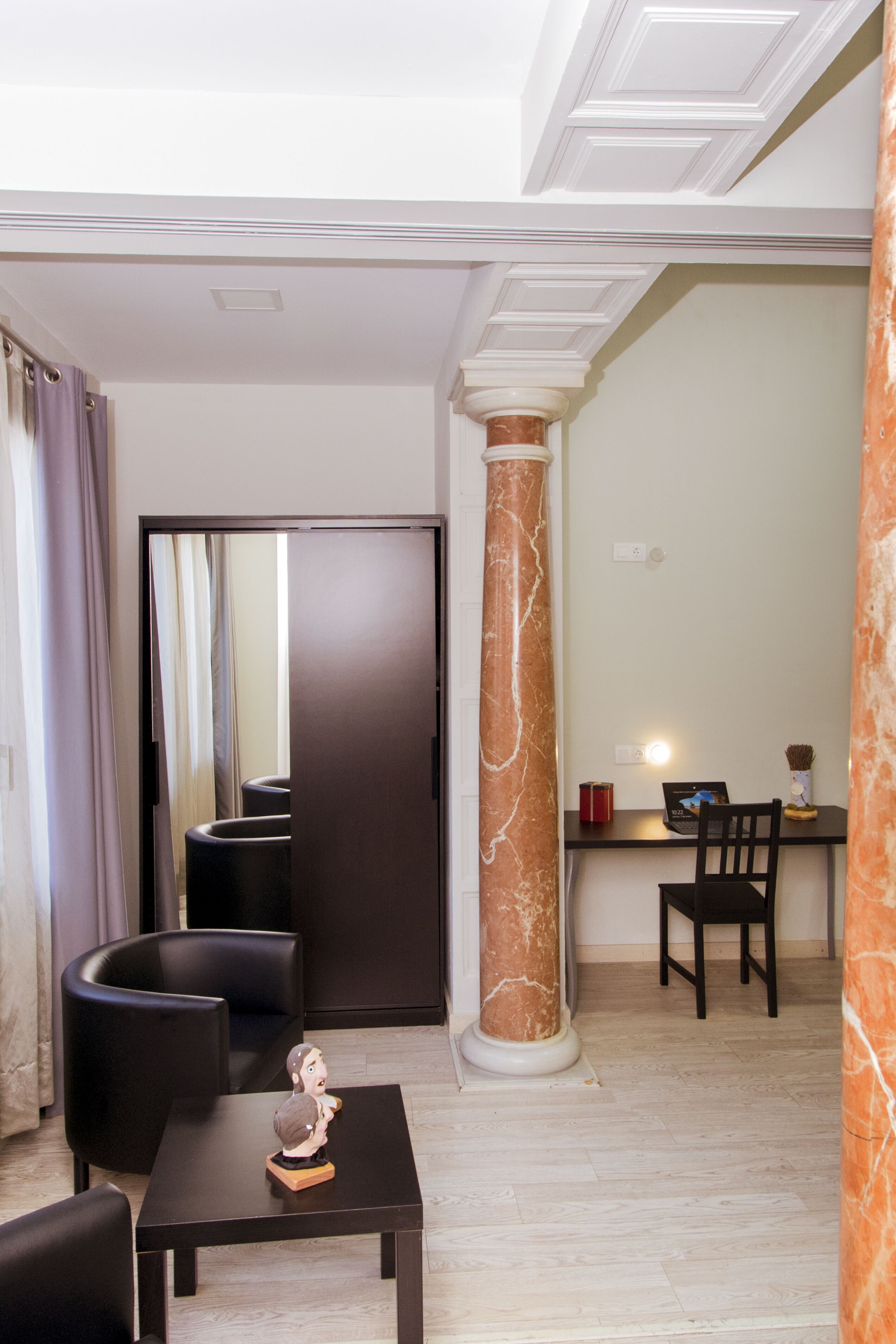 Habitación de hotel moderna con un escritorio de tonos oscuros, una pequeña estatua, una silla negra curvada, una columna de mármol grande y un espejo que refleja la habitación.