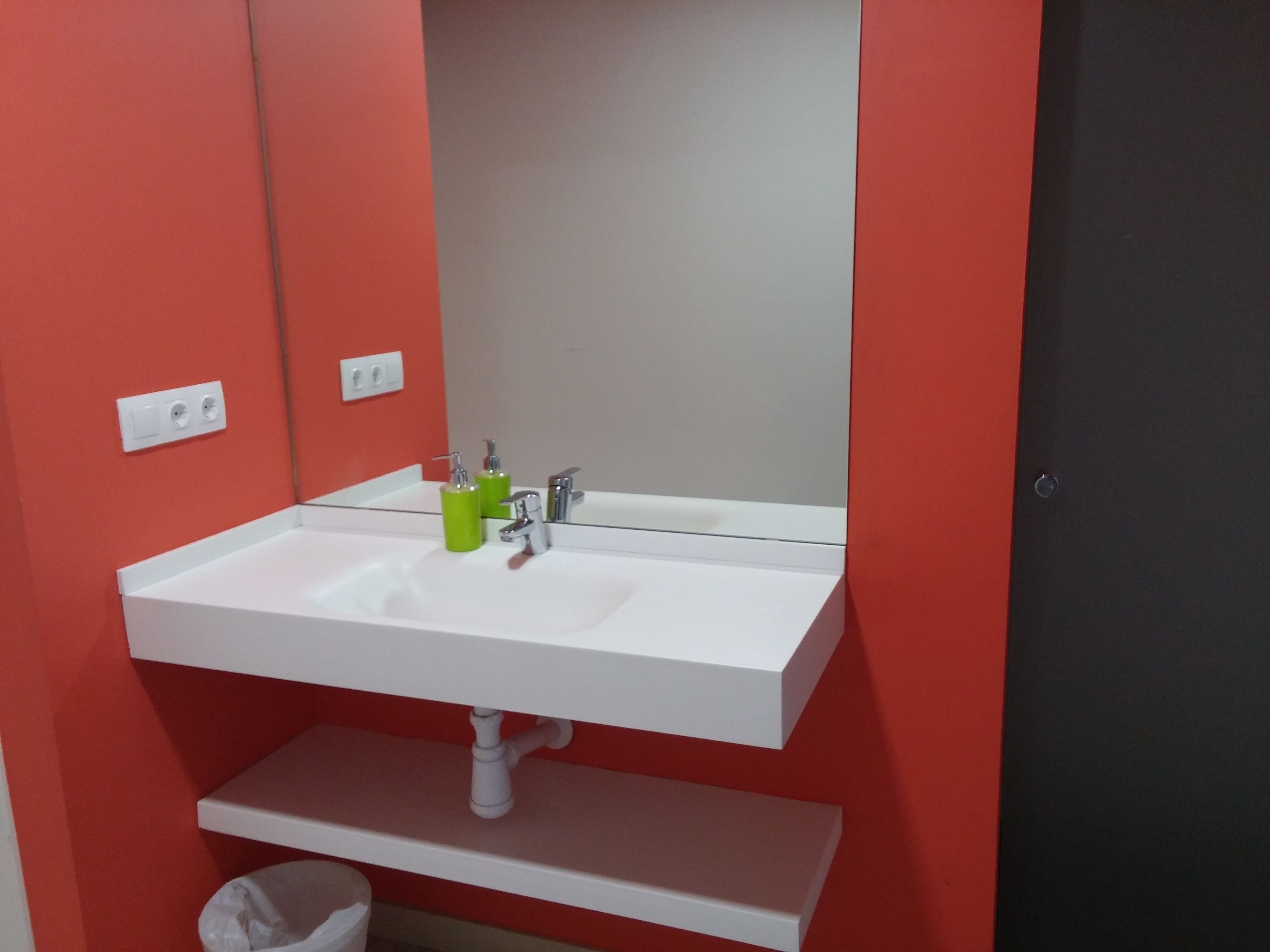 A white bathroom sink with a mirror above it, a green soap dispenser, and a white trash bin below.