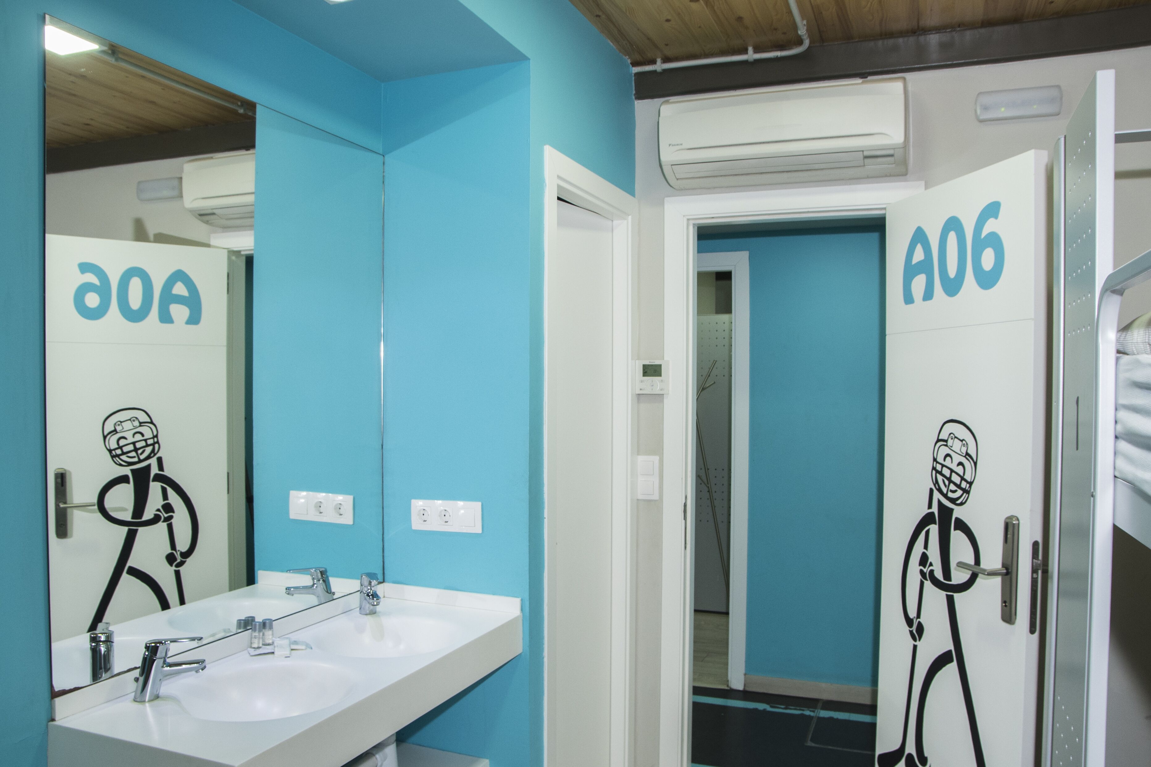 Un baño con pared azul, un lavamanos blanco y un espejo. Hay una puerta con la etiqueta "A06" y un personaje de dibujos animados.