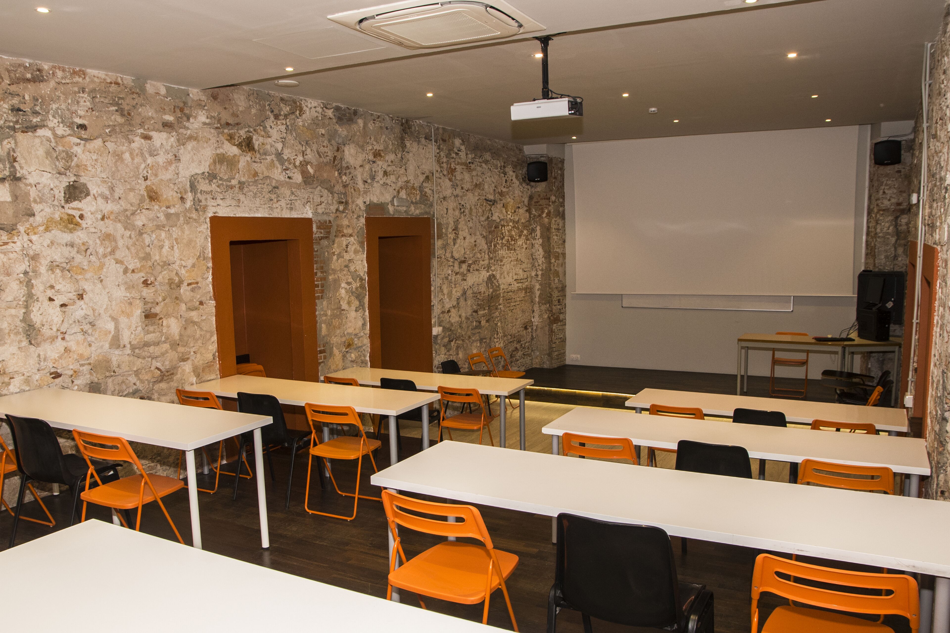 Une salle de conférence avec des tables blanches et des chaises orange, un écran de projection et un mur en pierre.