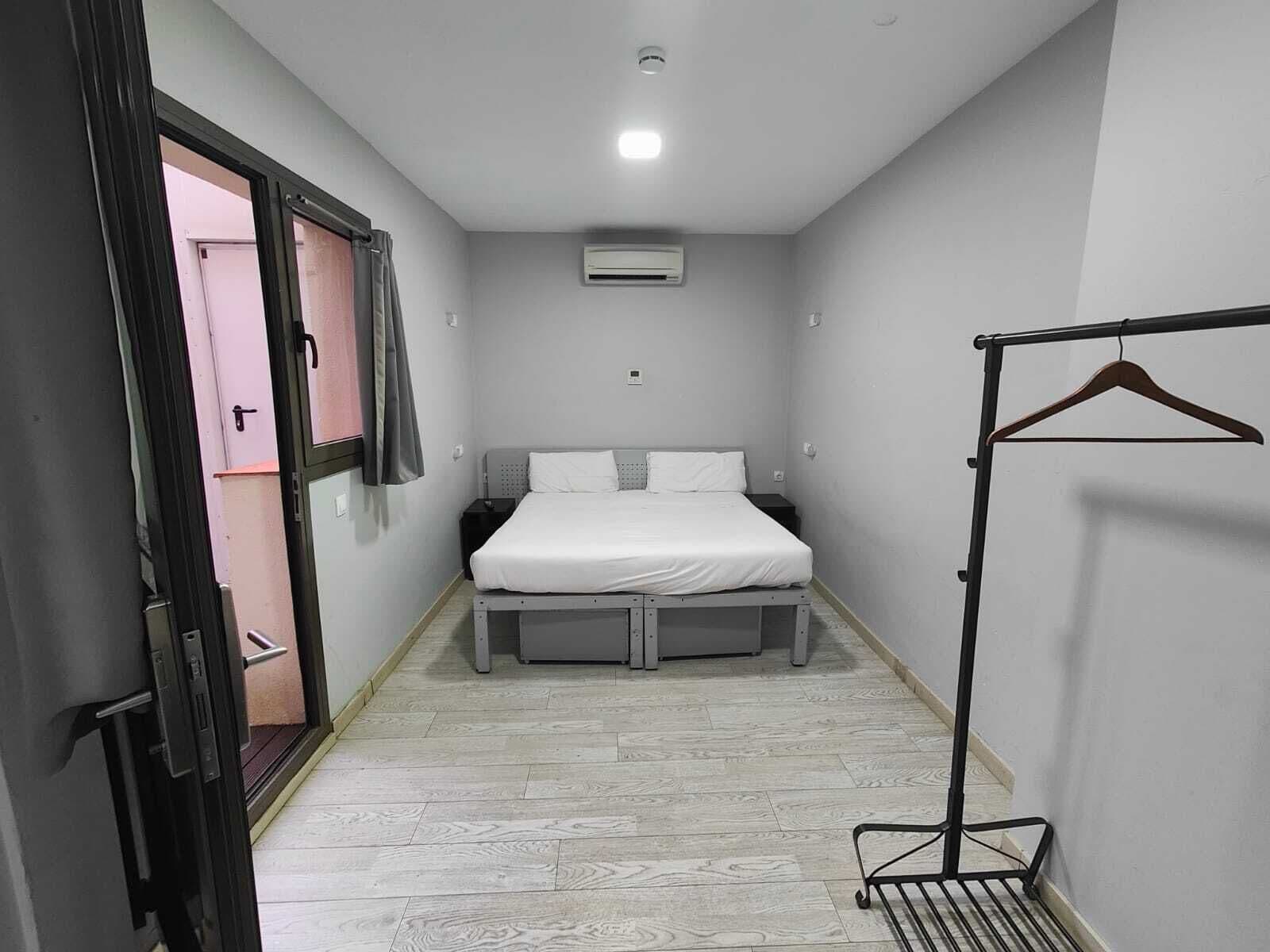 Une petite chambre d’hôtel étroite, avec un lit simple, une table de chevet, un porte-manteau mural et une porte donnant sur une autre pièce.