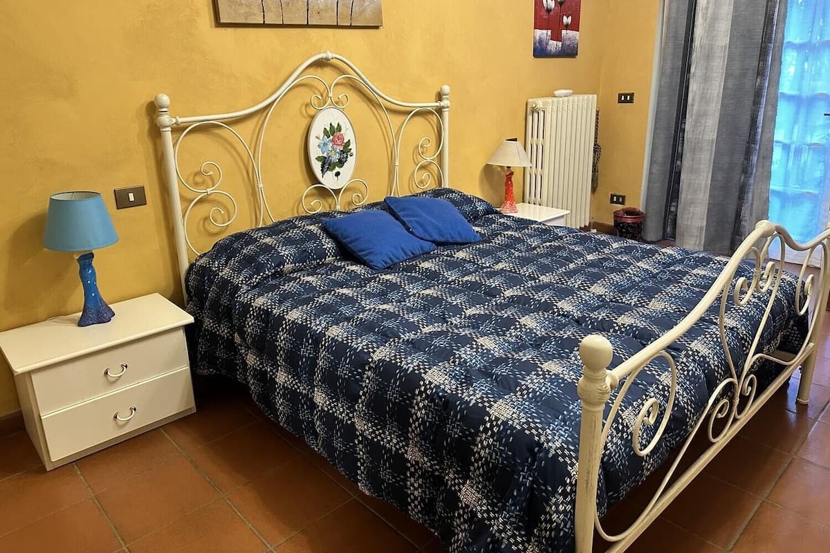 A bedroom with a bed, a nightstand, a lamp, a radiator, and artwork on the walls.