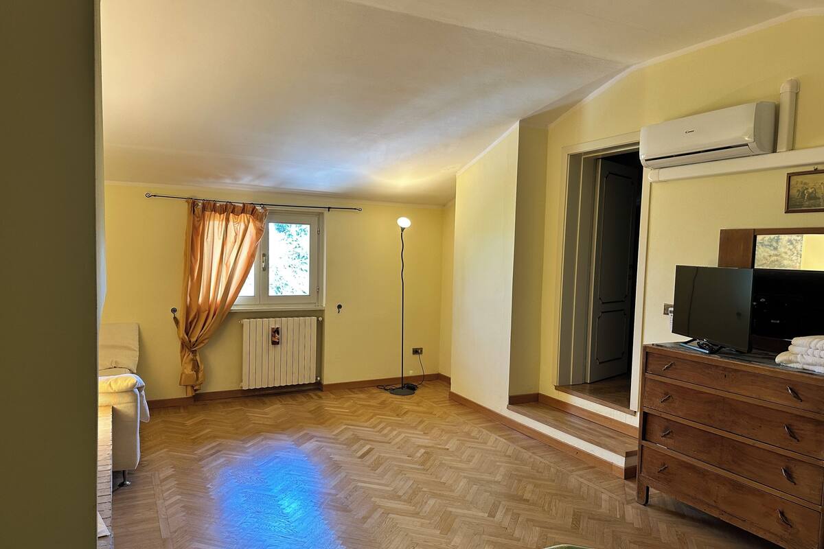 A room with a wooden herringbone floor, a radiator, a standing lamp, a television on a wooden cabinet, and a window with curtains.