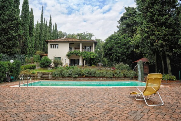 A pool area with a lounge chair, a house with a balcony, and a garden with trees.