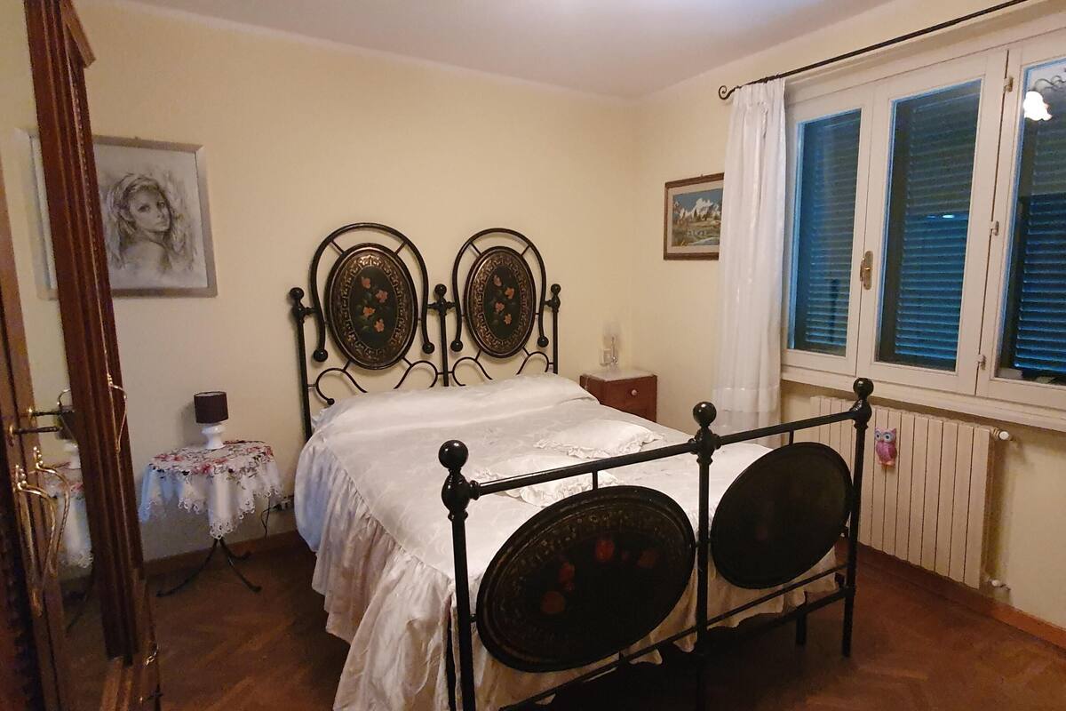 A bedroom with a four-poster bed, a small table with a lamp, and a window with shutters.
