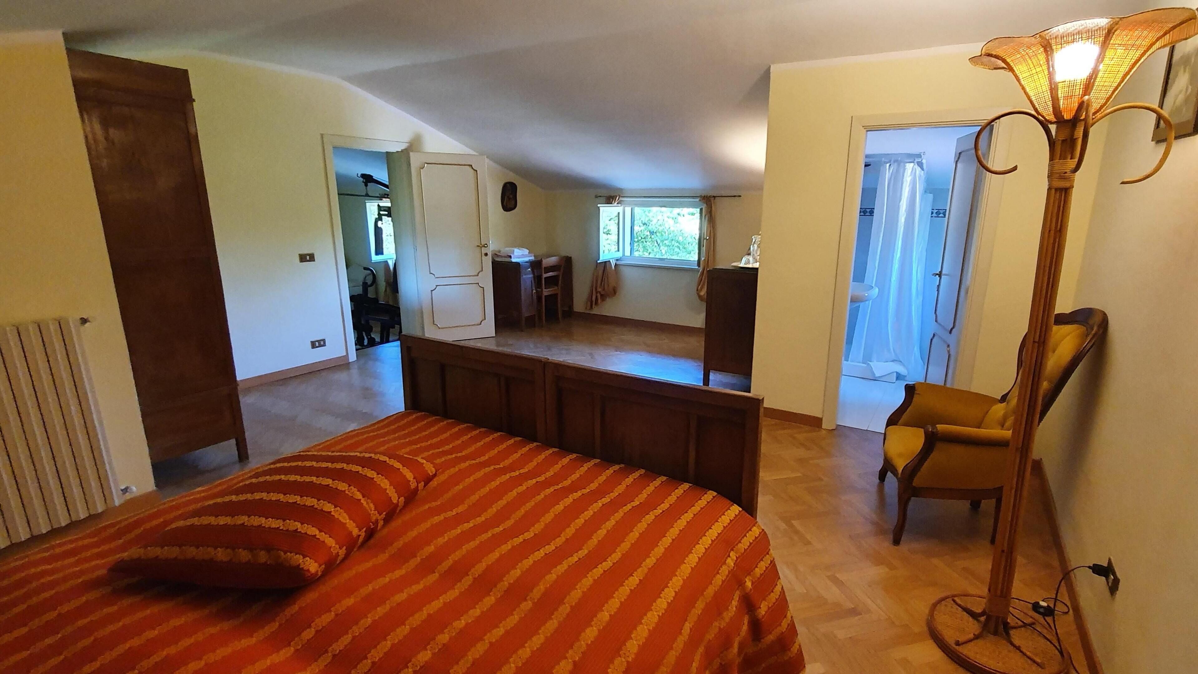 A bedroom with a bed, a desk, and a chair. There is a window with curtains and a radiator.