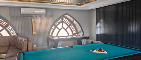 A pool table with a teal felt surface, a brown leather sofa, and a decorative arched window with a geometric pattern.