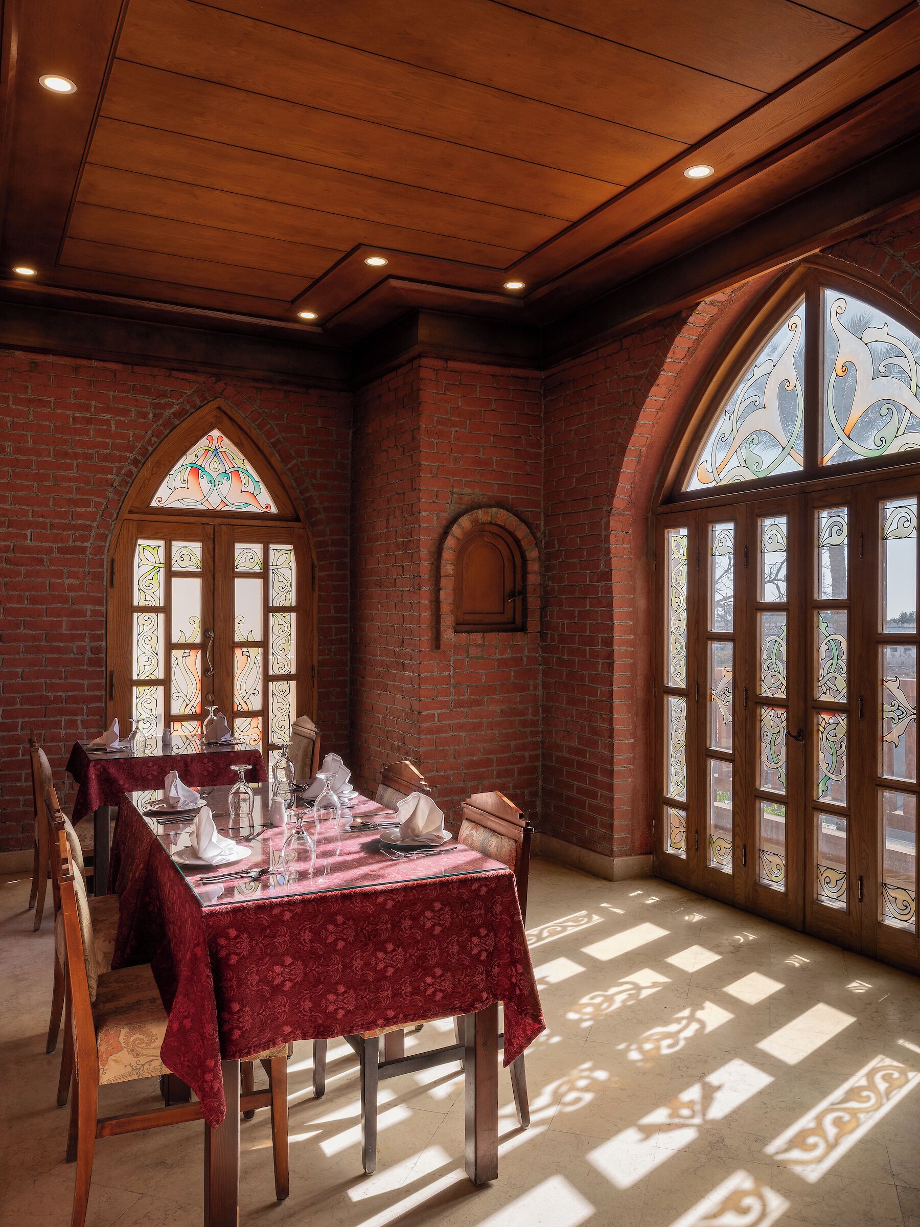 Comedor con pared de ladrillo, techo de madera y ventanas con vitrales. Las mesas están preparadas con manteles blancos y cubiertos.