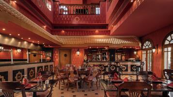 A restaurant with a traditional interior, featuring a central dining area with multiple tables and chairs, a bar area with a counter, and a decorative ceiling.