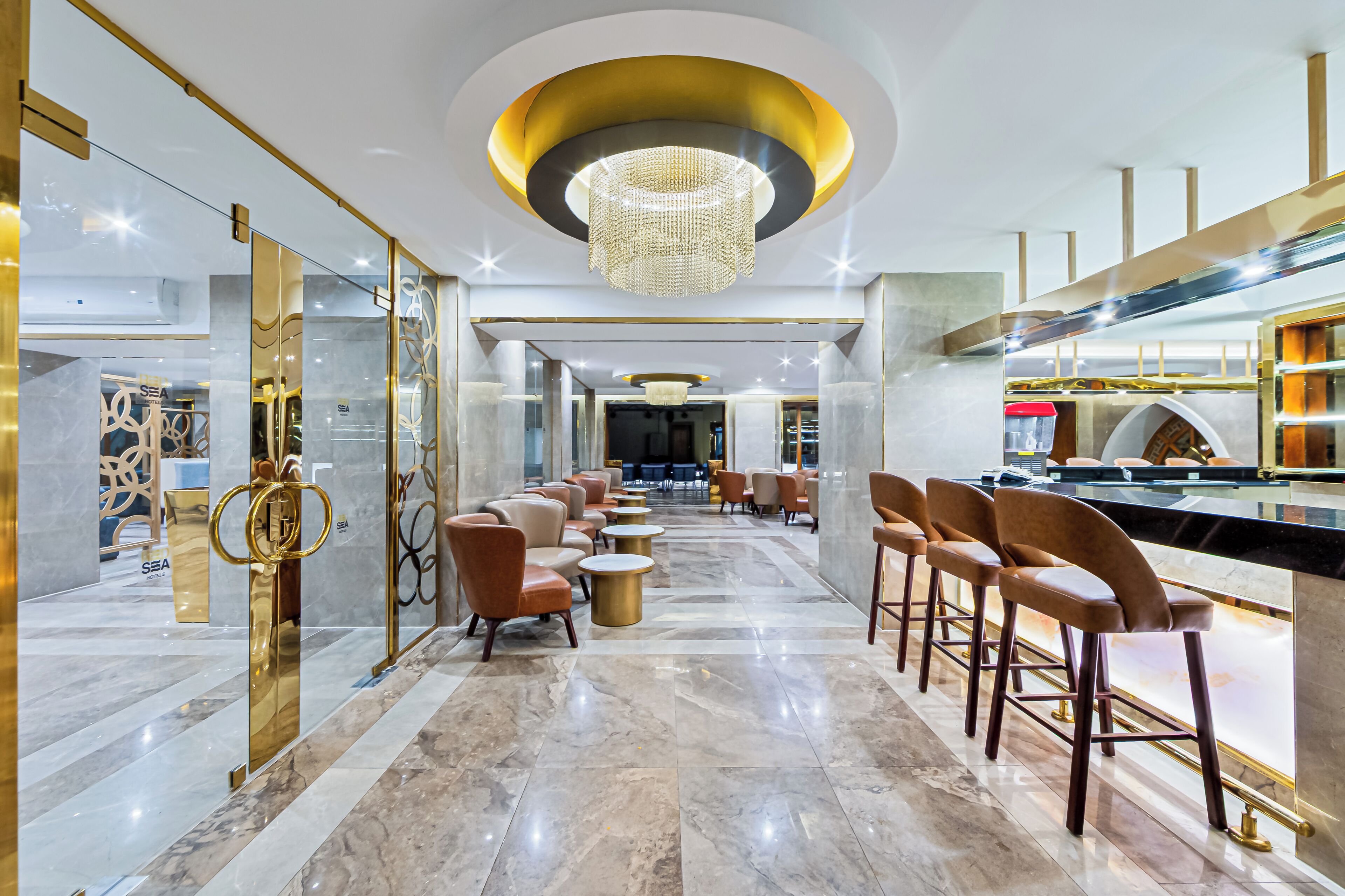 A modern hotel lobby with a marble floor, a central chandelier, and a bar area with stools.