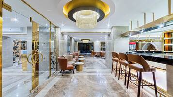 A modern hotel lobby with a marble floor, a central chandelier, and a bar area with stools.
