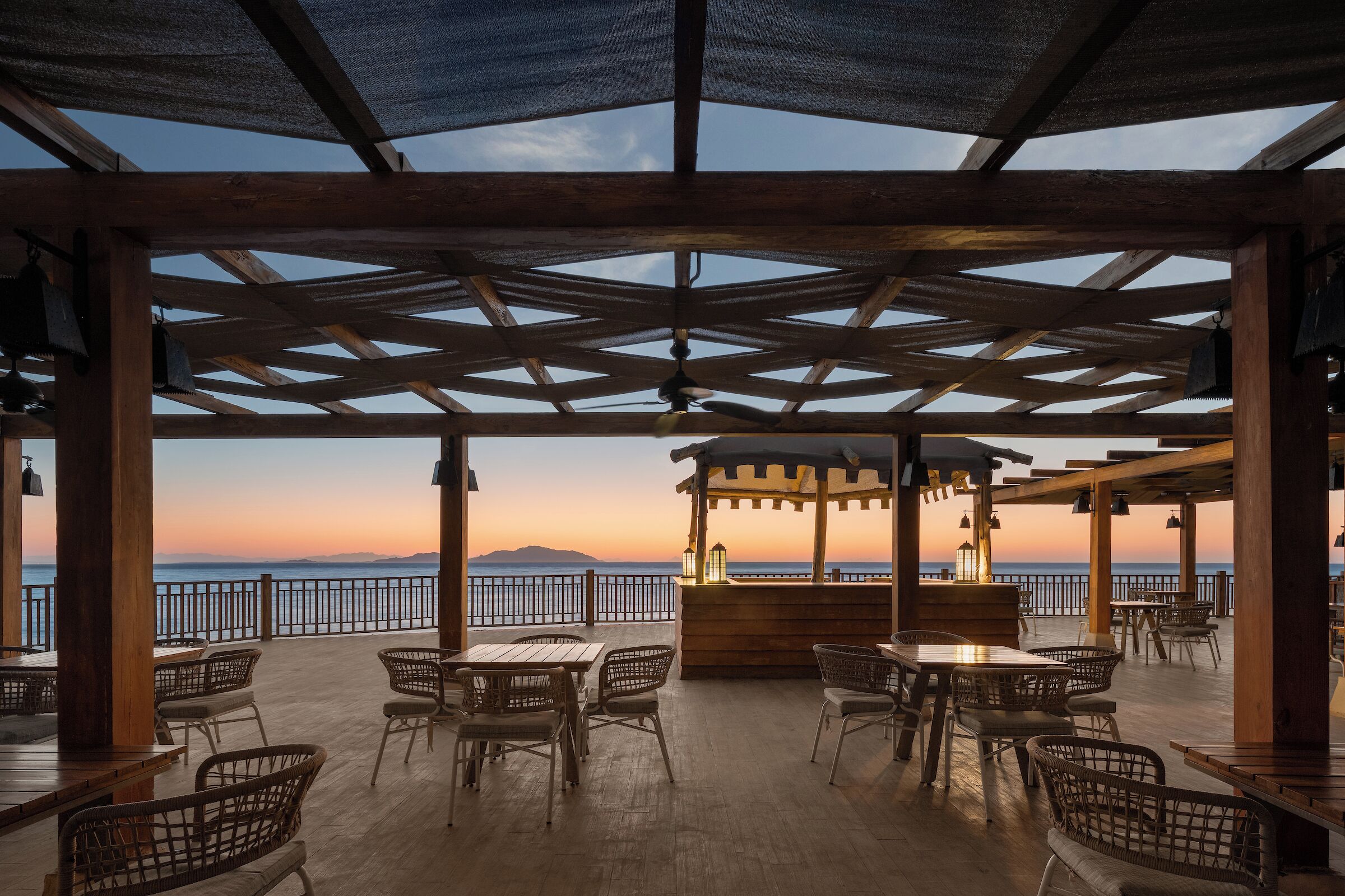 An outdoor patio with wooden tables and chairs, a bar area, and a view of the sea at sunset.