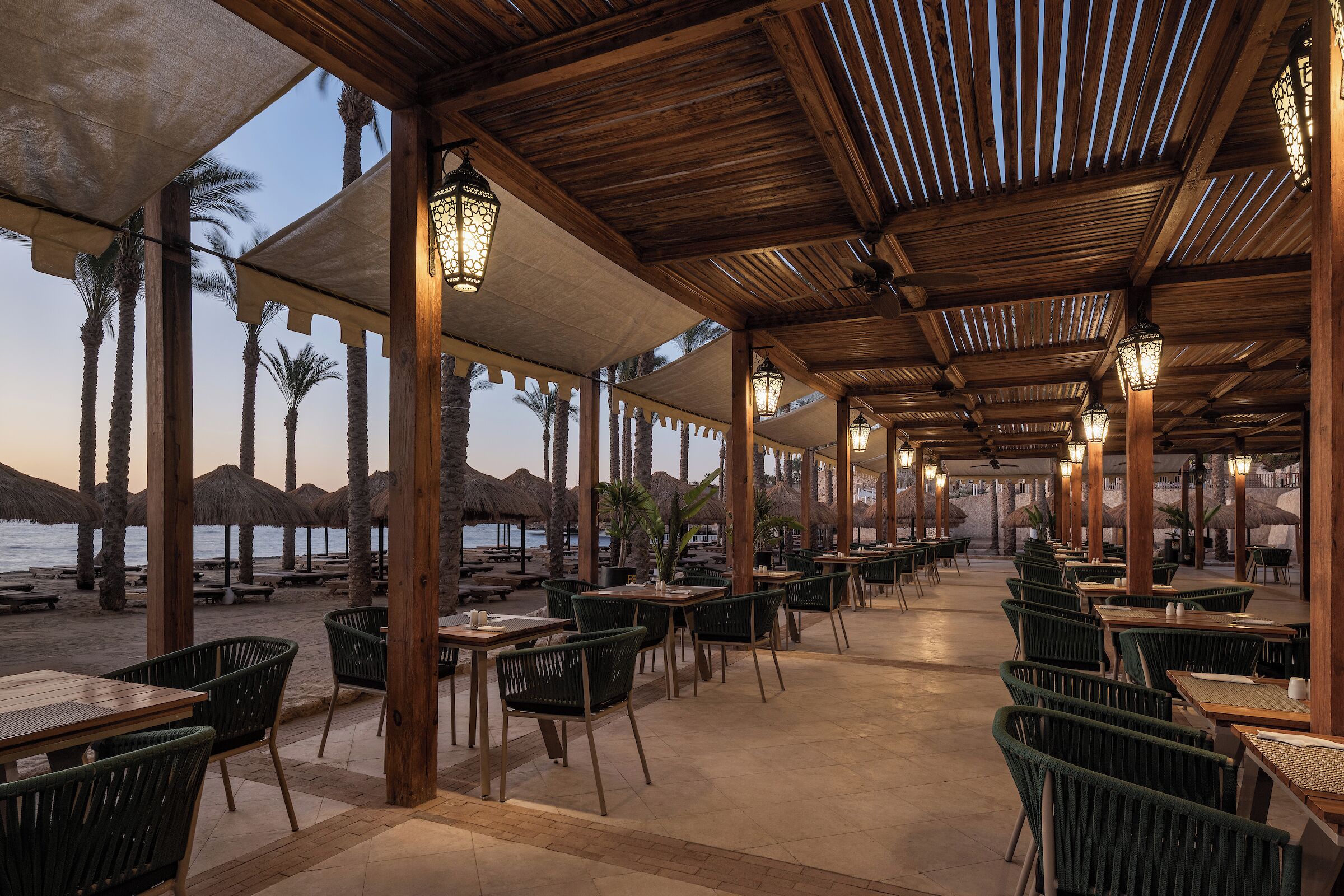 A covered outdoor seating area with tables and chairs, palm trees, and a view of the beach.
