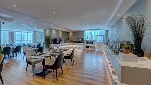 Een ruime hotel eetzaal met meerdere tafels en stoelen, een centraal eiland met keukenapparatuur, en grote ramen die voor natuurlijk licht zorgen.