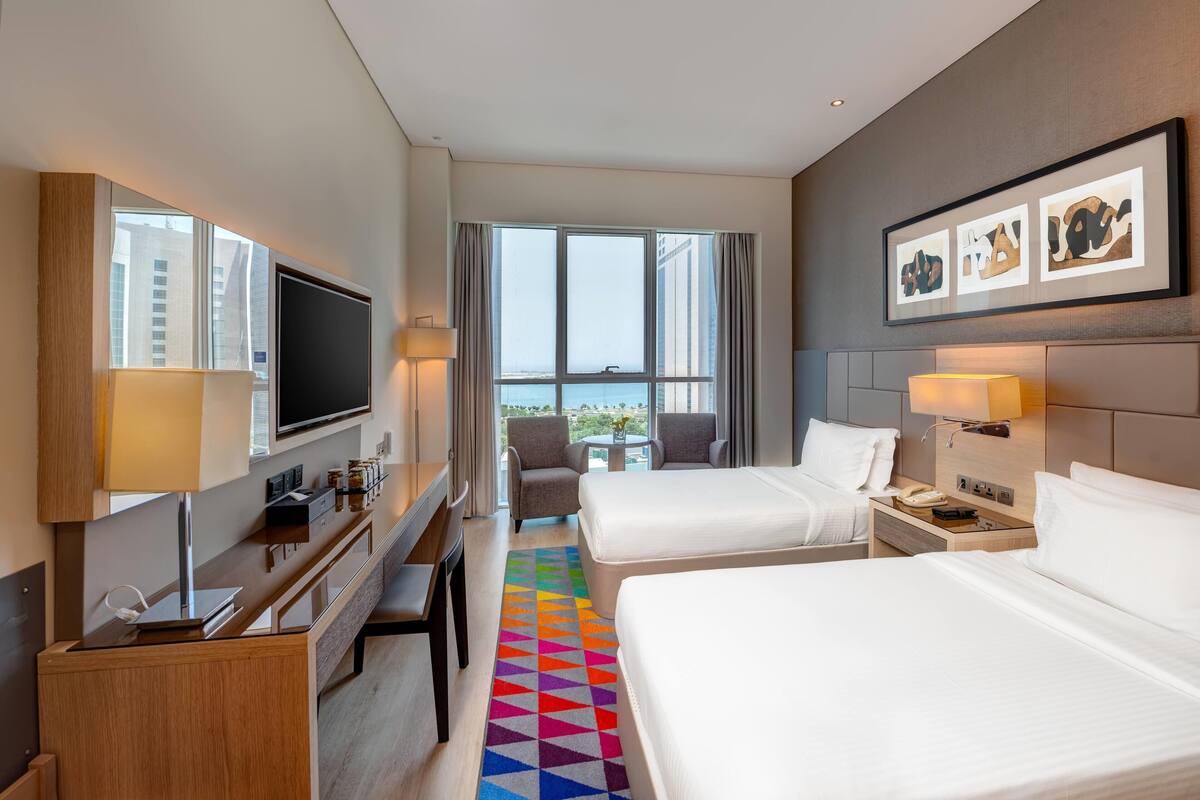 A hotel room with two beds, a desk, a TV, and a view of the city.