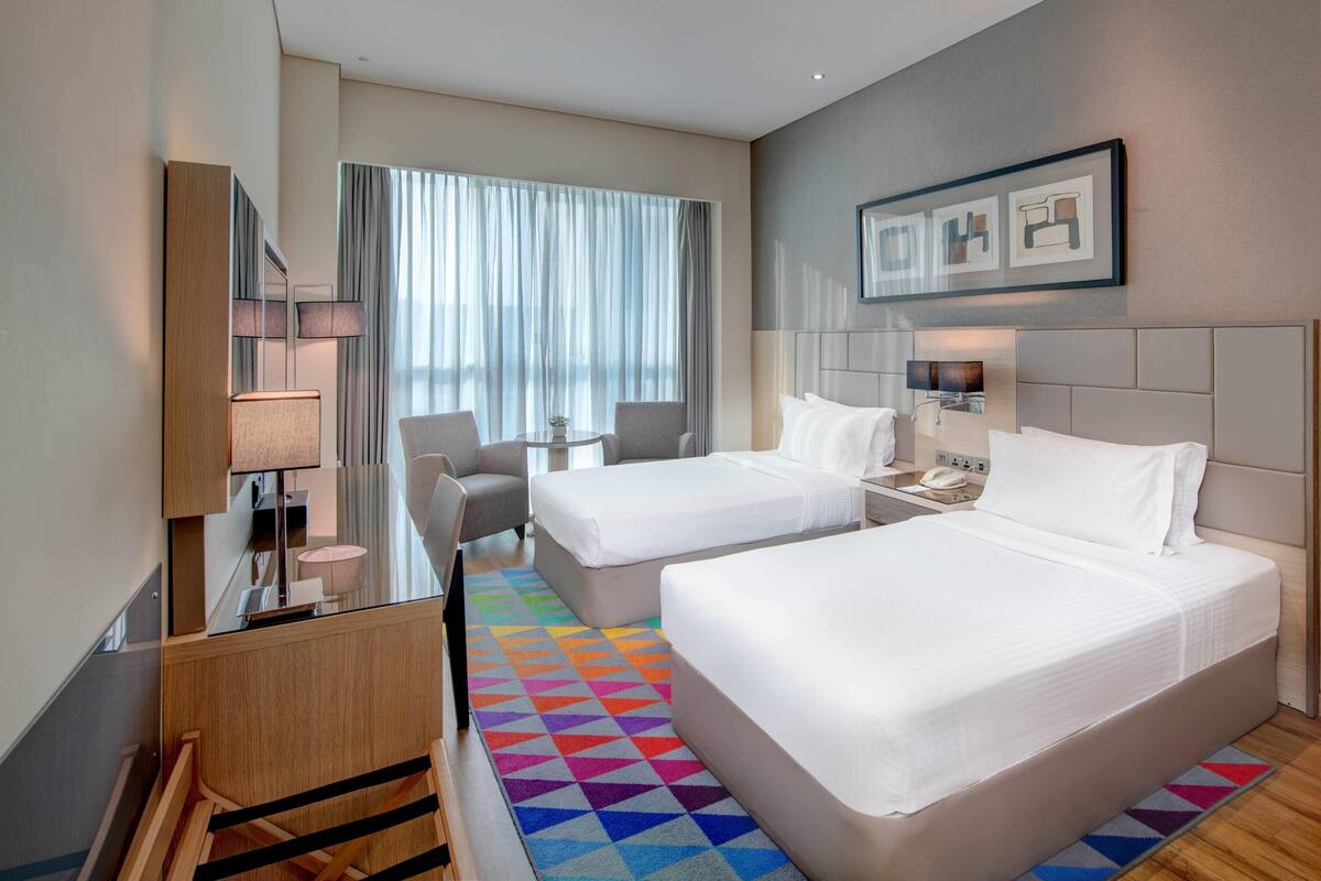 A hotel room with two beds, a desk, a chair, a lamp, and a large window with curtains.