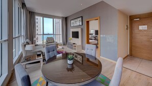 Een moderne hotelkamer met een ronde eettafel, grijze banken, een televisie en een groot raam met uitzicht op de stad.