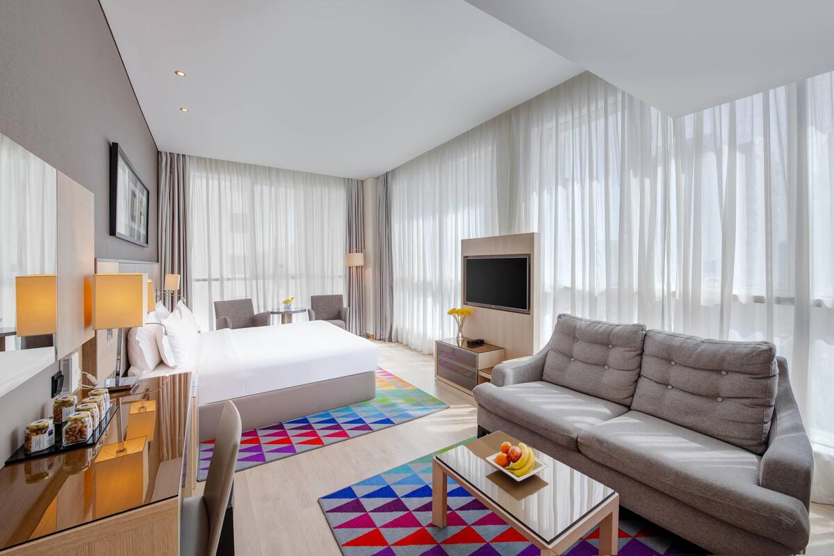 A modern hotel room with a large bed, a sofa, a TV, and a desk with lamps and a fruit bowl.