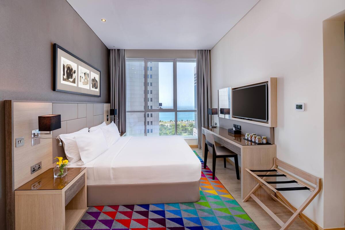 A hotel room with a large bed, a desk, a TV, and a view of the city.