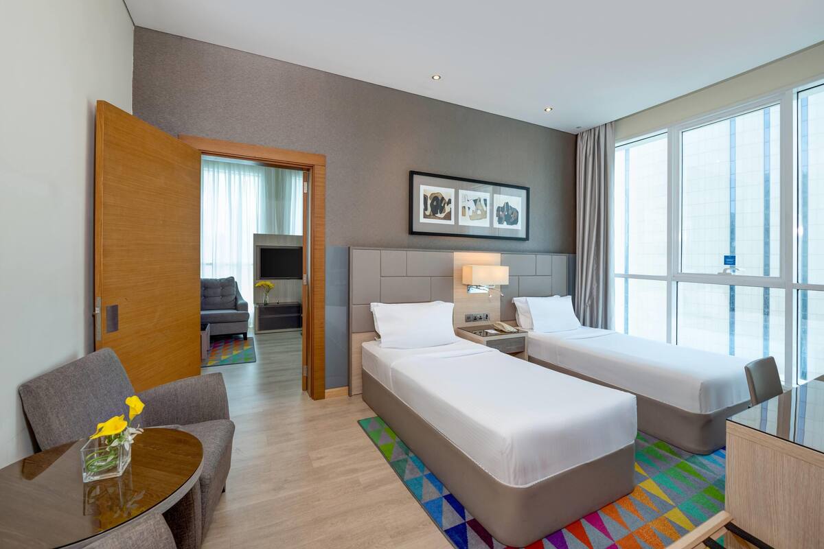 A hotel room with two beds, a desk, a chair, a small table with flowers, and a colorful rug.