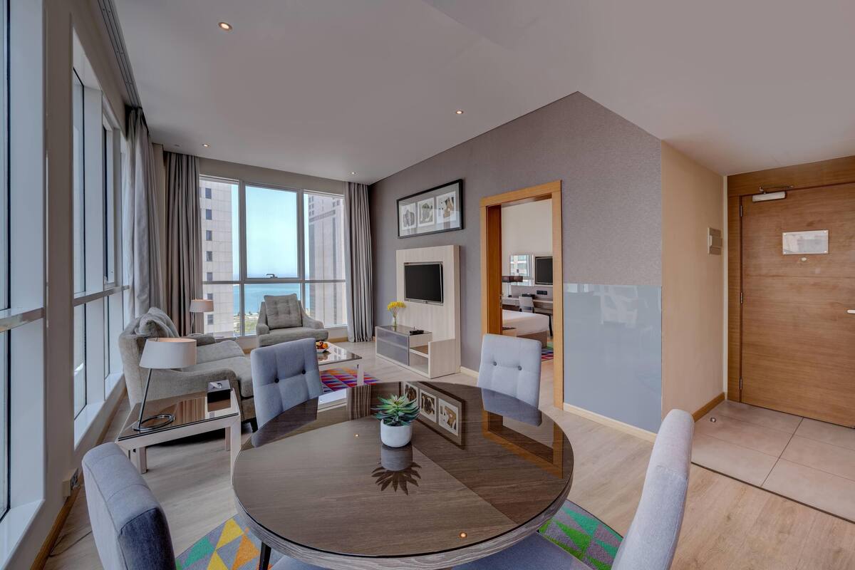 A modern hotel room with a round dining table, grey sofas, a TV, and a large window offering a city view.