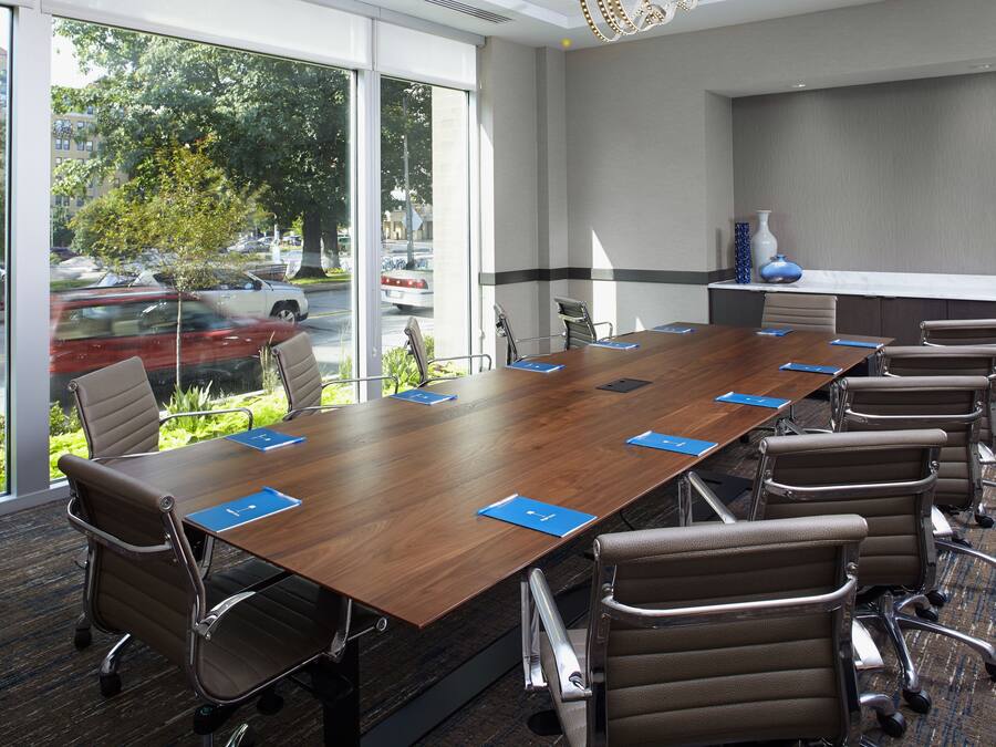 A conference room with a long wooden table, multiple chairs, and large windows offering a view of parked cars and trees outside.