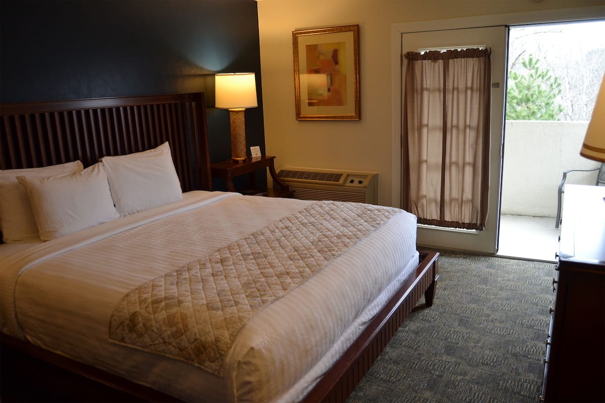 A hotel room with a large bed, a nightstand with a lamp, a painting on the wall, and a view through a sliding door.