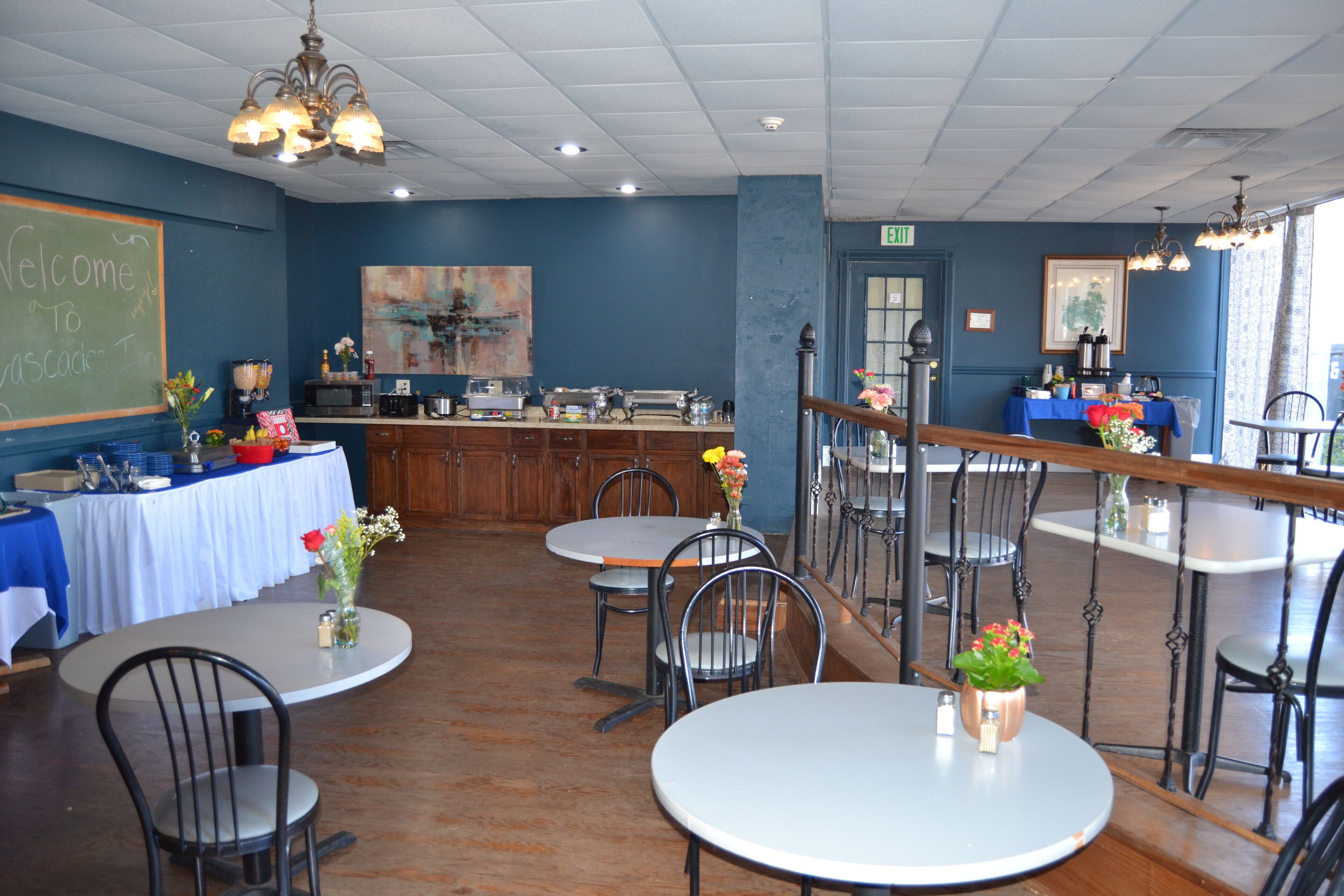 A restaurant interior with round tables, chairs, a chalkboard sign, artwork, and a bar area.