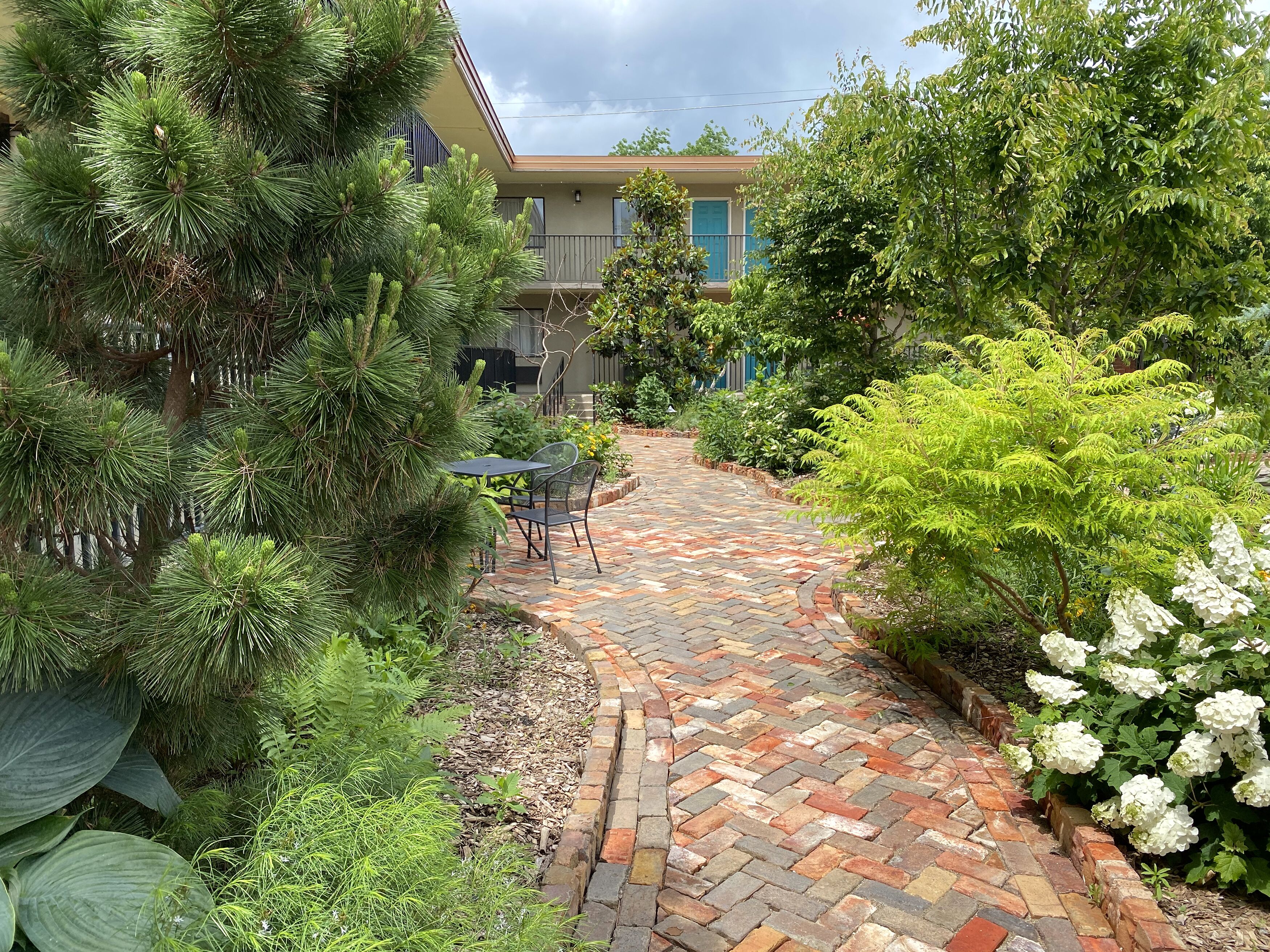 A brick pathway leading to a patio area with a table and chairs, surrounded by lush greenery and flowering plants.