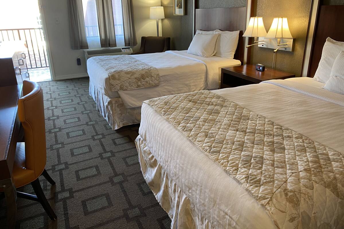 A hotel room with two beds, a desk, and a chair. There are two bedside lamps and a window with curtains.