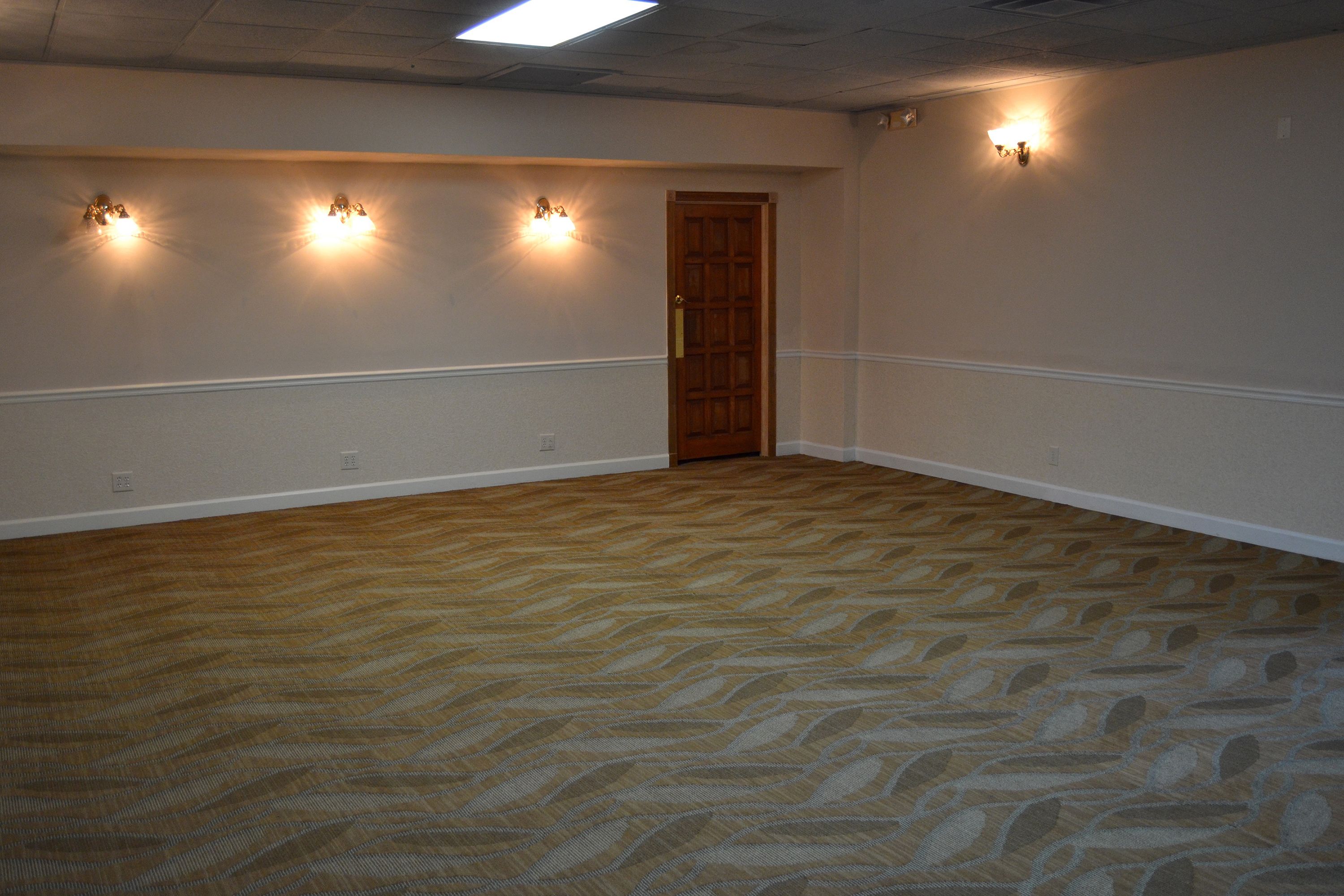 A spacious room with a patterned carpet, a wooden door, and wall-mounted lighting fixtures.