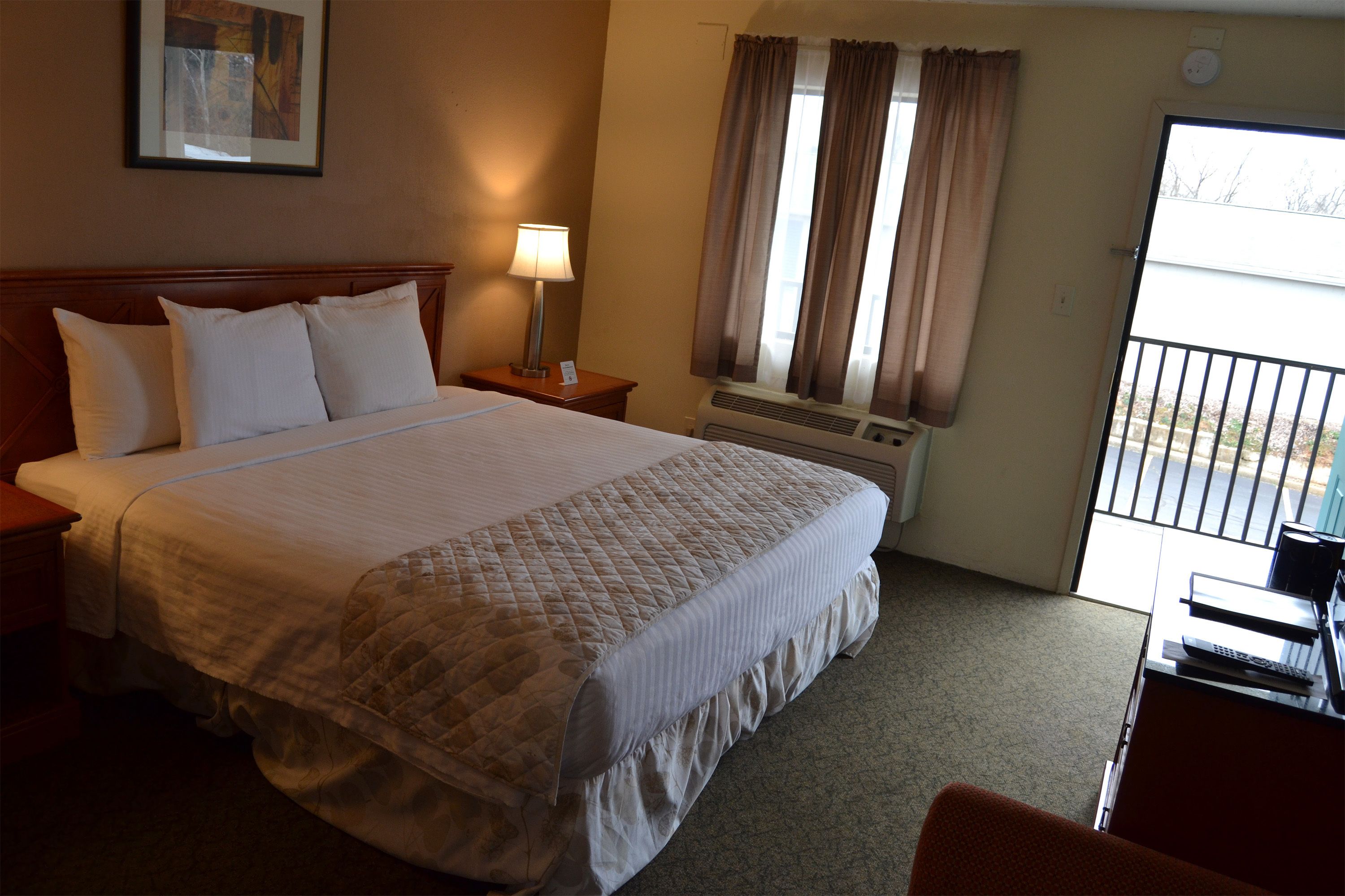 A hotel room with a large bed, a nightstand, a lamp, a window with curtains, and a view of the outside.
