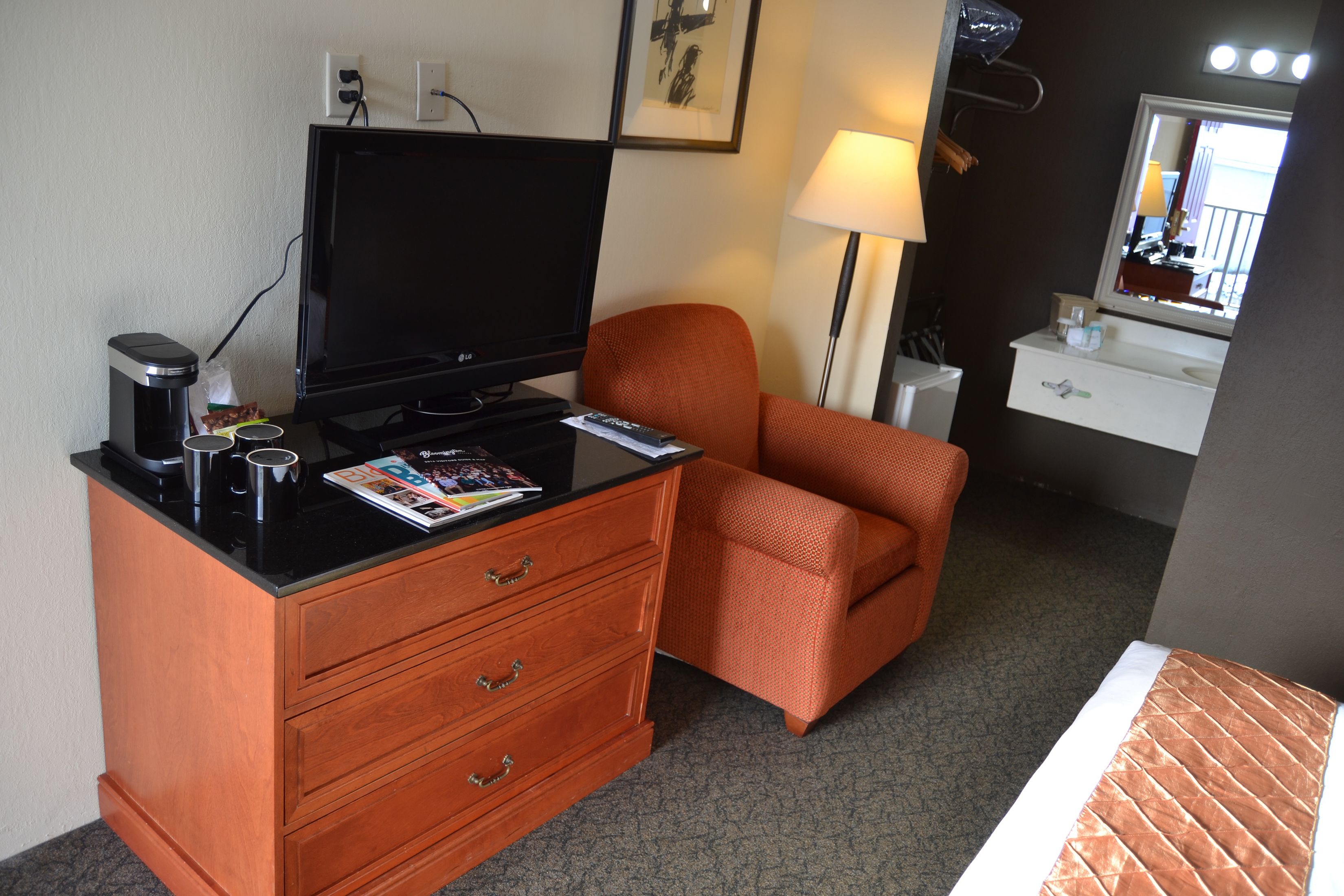 Une chambre d’hôtel avec une commode en bois, un téléviseur, une lampe et une salle de bain avec un miroir.