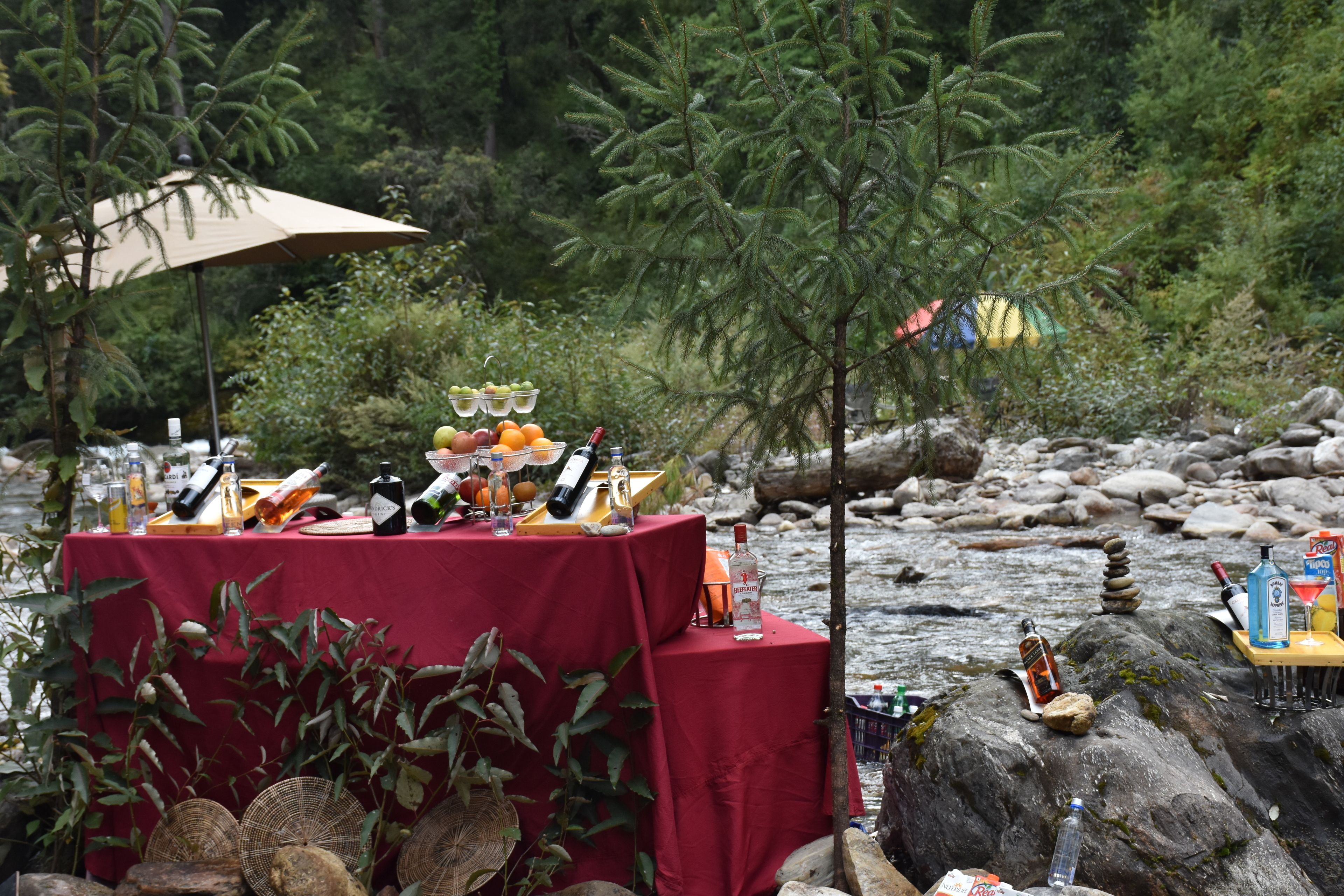 A table set up outdoors with a red tablecloth, various bottles, glasses, and fruits.