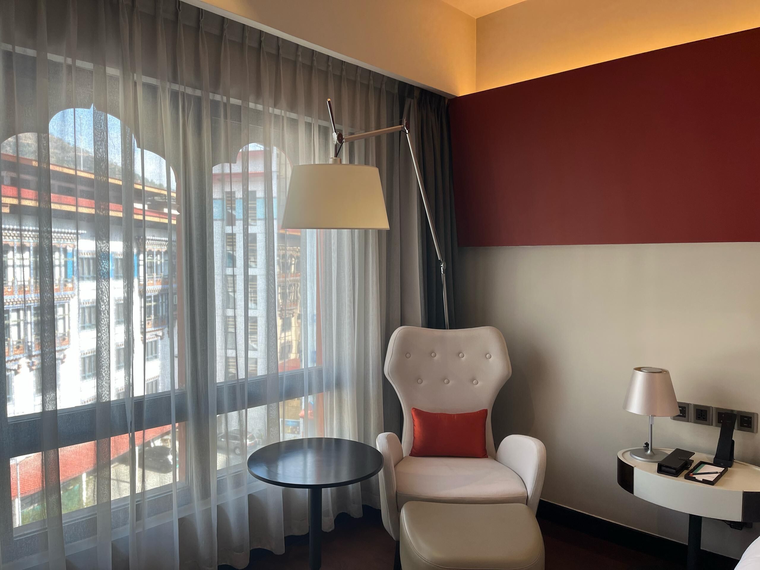 A hotel room with a view of a cityscape through sheer curtains, a white upholstered chair with a red cushion, a small round table, and a desk with a lamp and a phone.