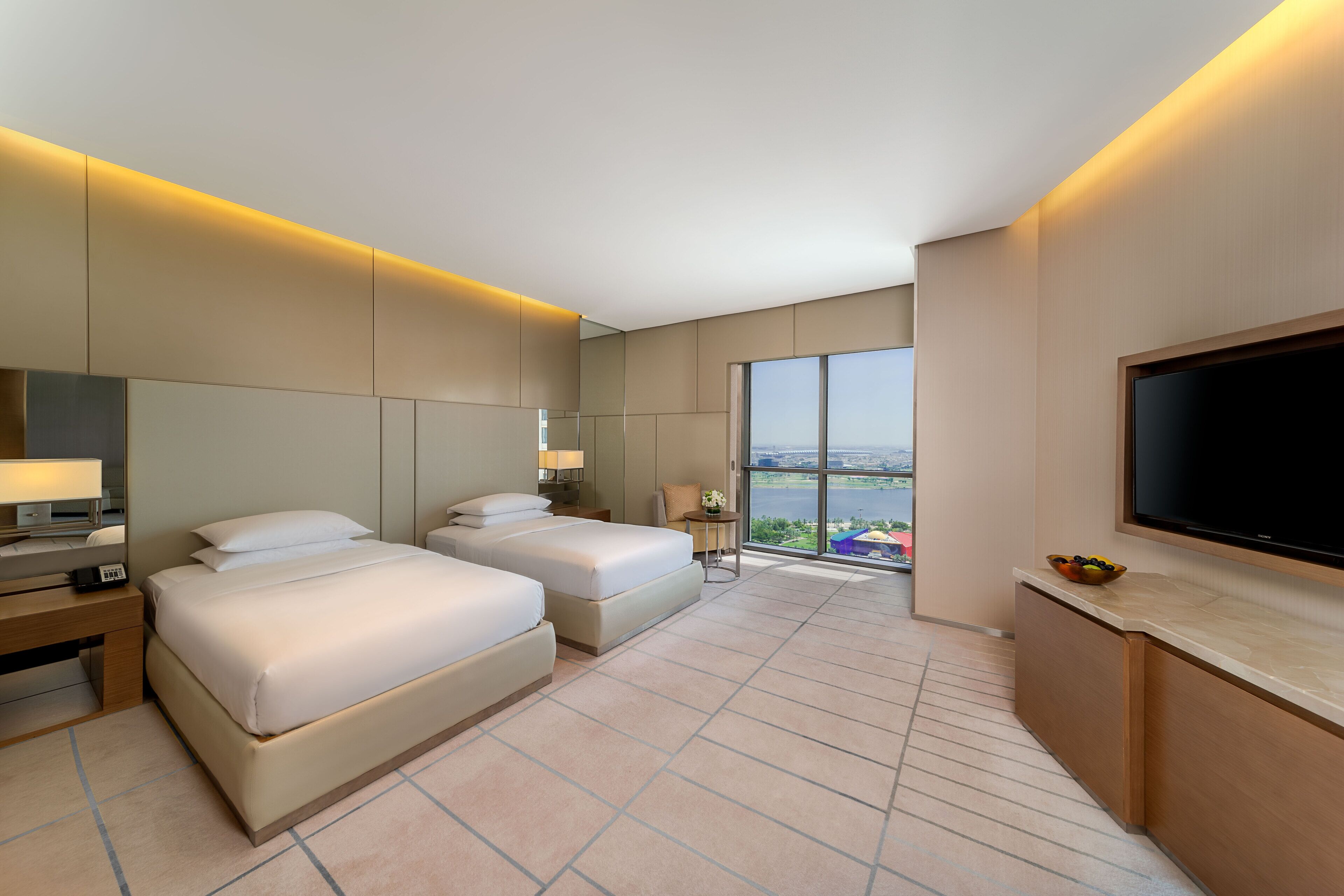 Quarto de hotel moderno com duas camas, televisão de tela plana, escrivaninha com luminária e vista para a cidade através de grandes janelas.