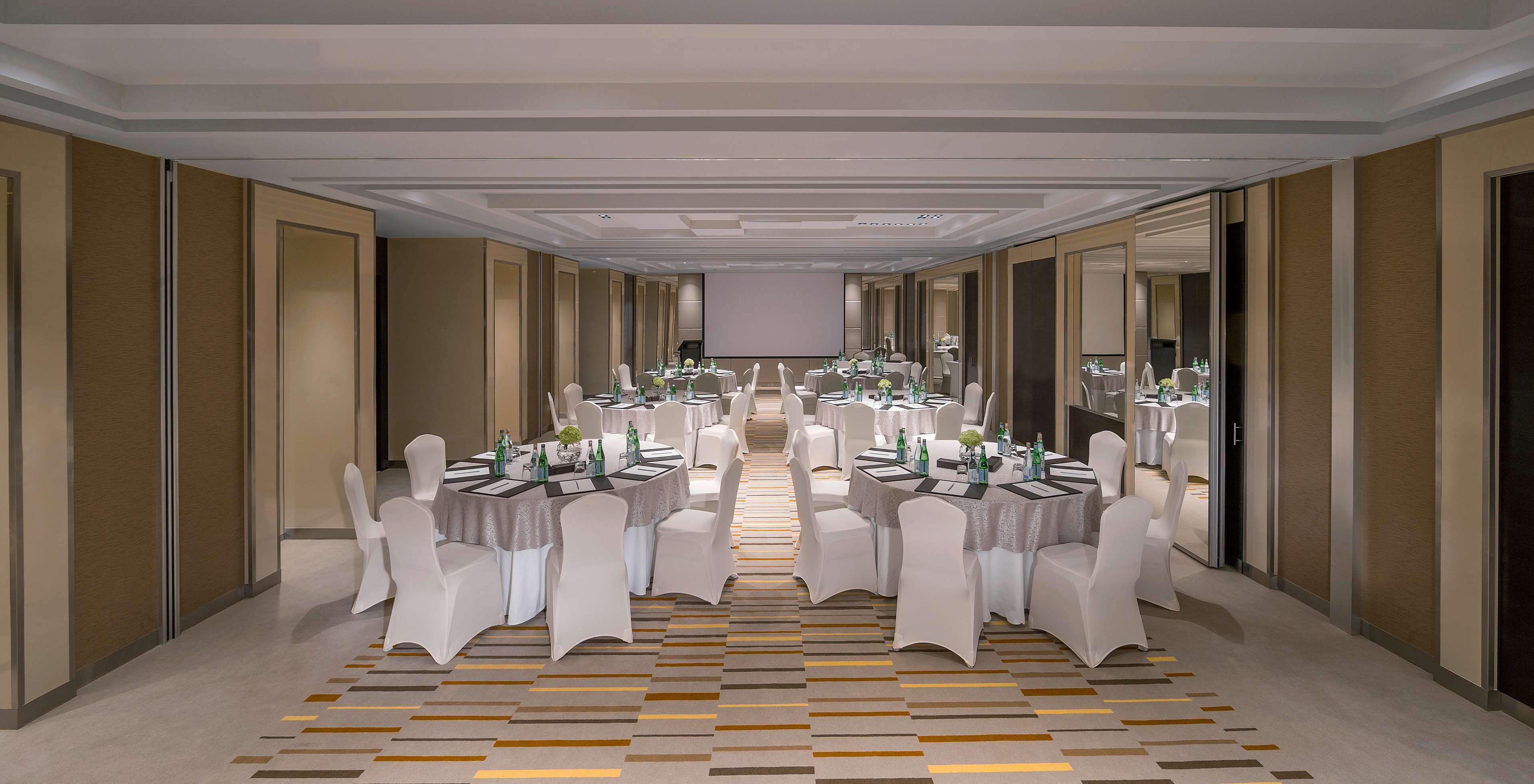 A long, narrow hotel corridor with multiple round tables and white chairs, each set with tablecloths and centerpieces.