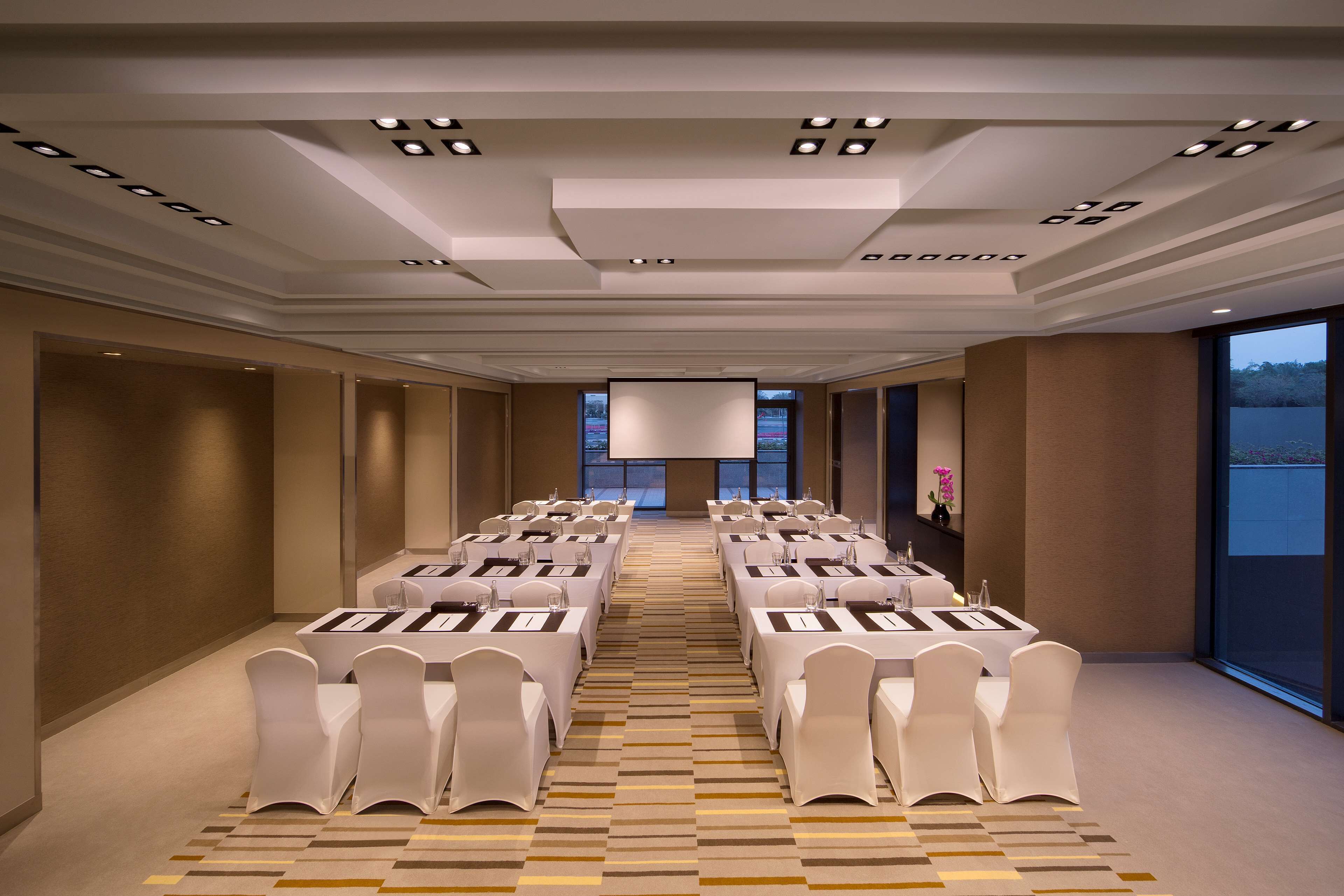 A conference room with tables set for a meeting, a large screen at the front, and a view of the outdoors through large windows.