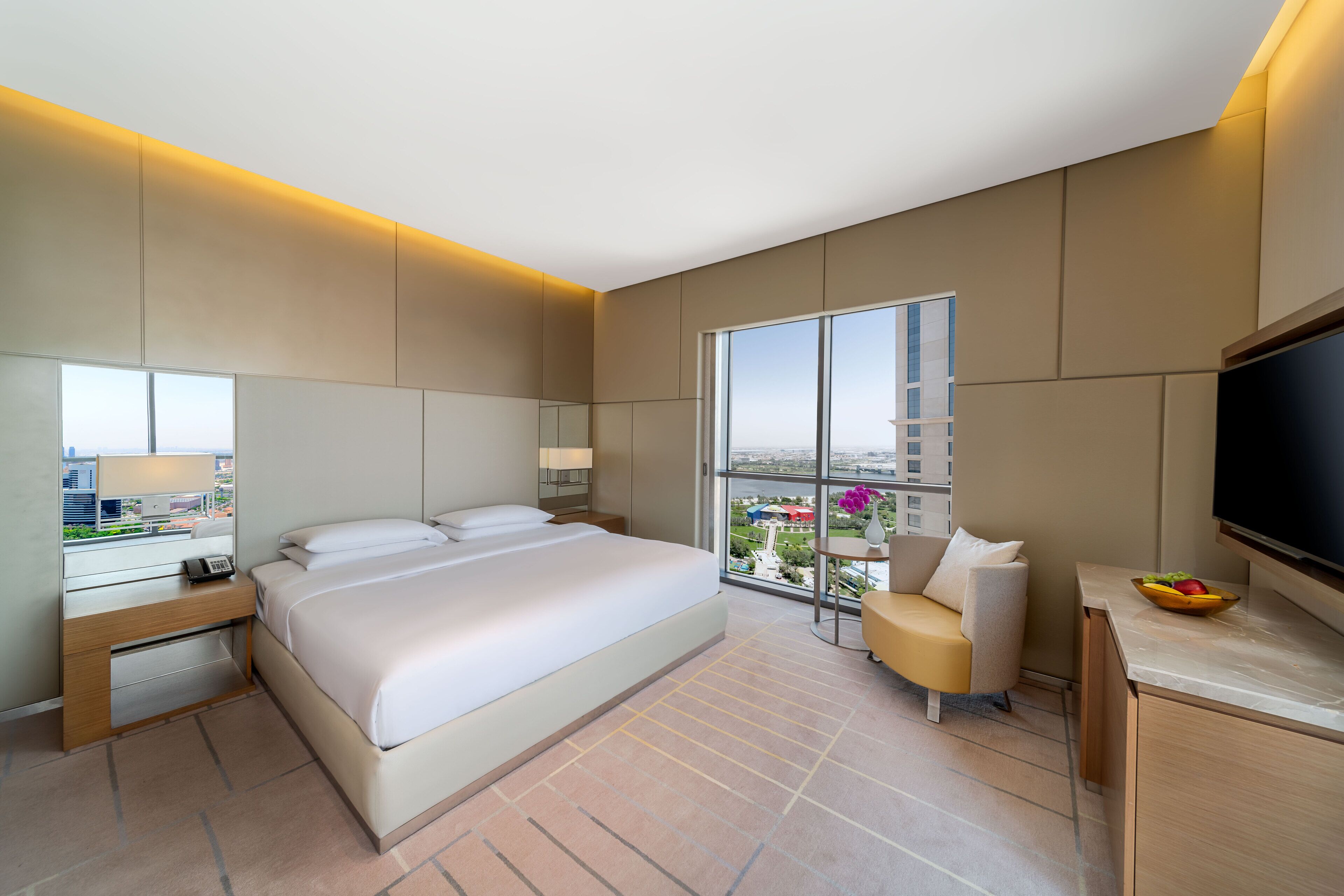 A modern hotel room with a large bed, a desk, a chair, a TV, and a large window offering a city view.