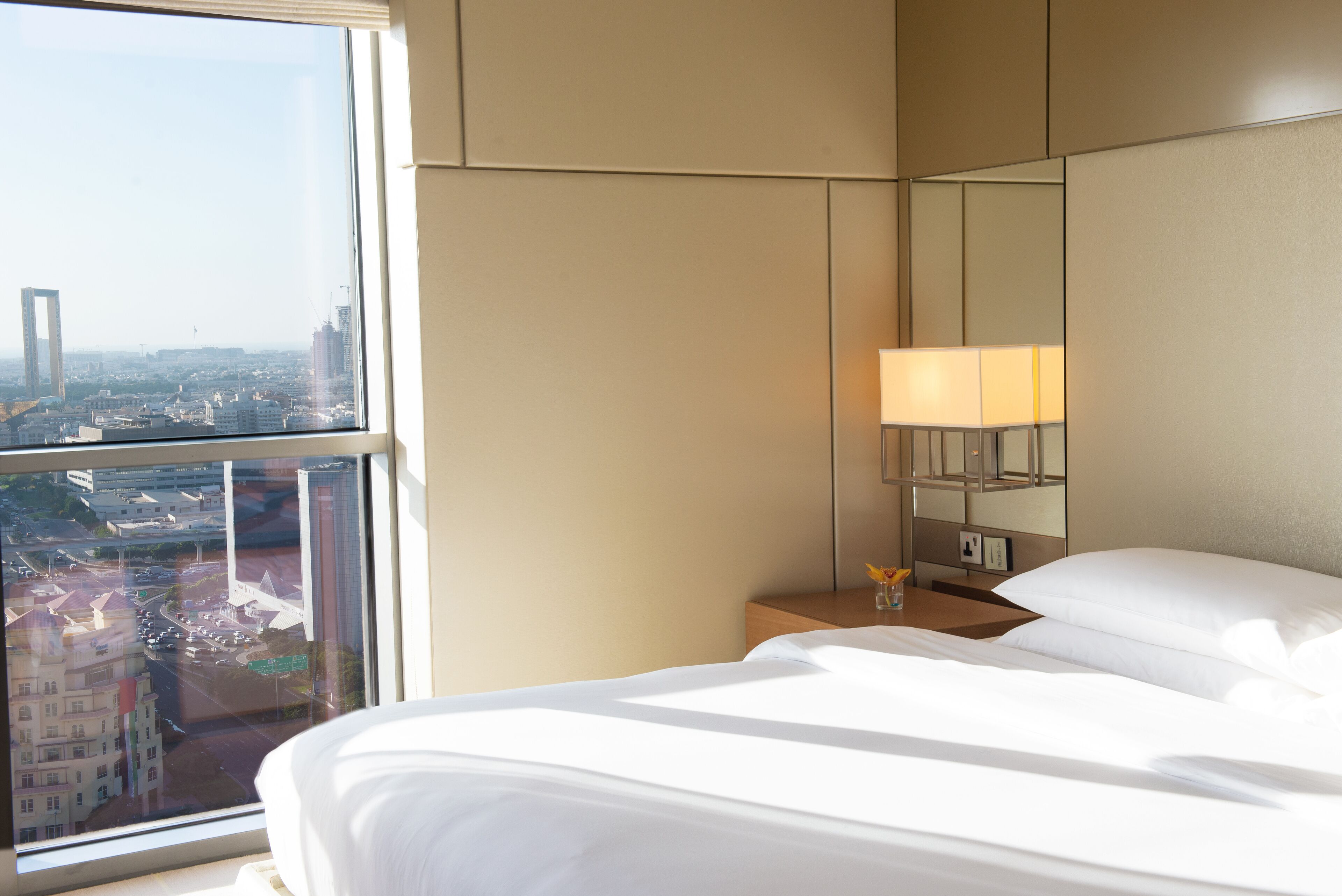 A hotel room with a large window, a bed, a bedside table with a lamp, and a mirror.